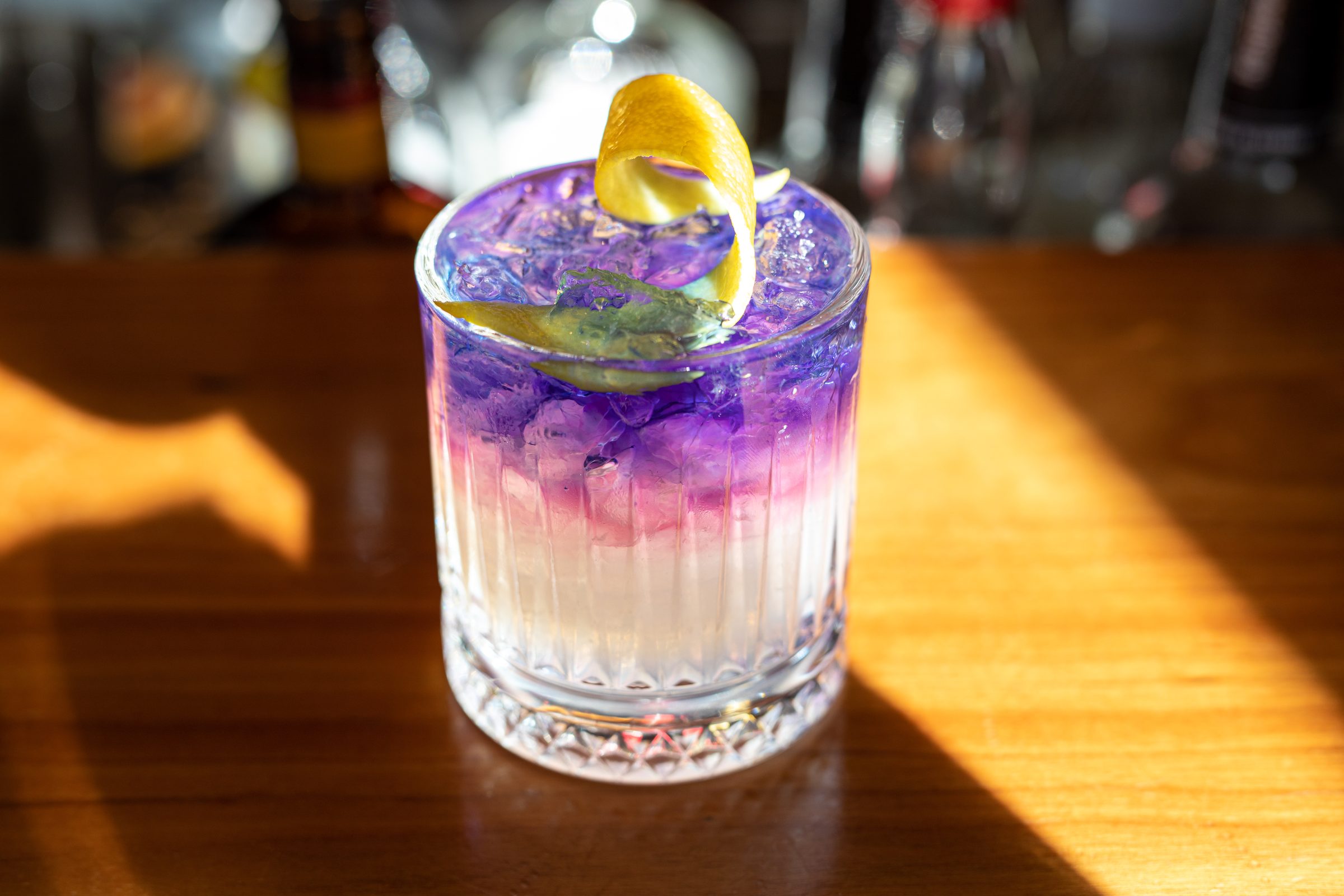 Violett und weiß geschichteter Cocktail mit Zitronenzeste im Kristallglas auf dem sonnigen Holztresen des Ludwig in Mosbach