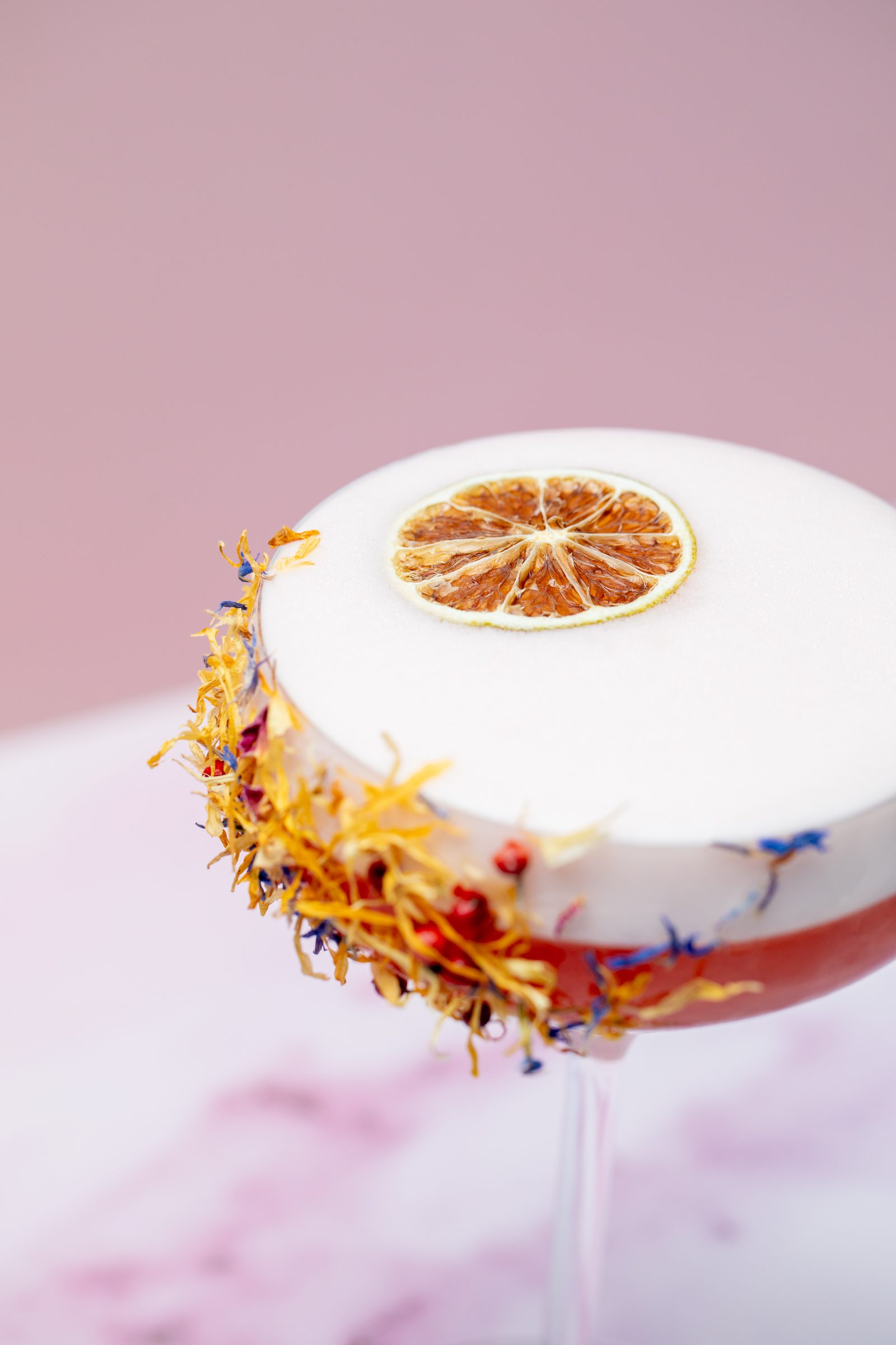 Cocktail mit weißer Schaumkrone, getrockneter Orangenscheibe und Blütenrand vor rosa Hintergrund, Cocktailfoto für das Ludwig in Mosbach