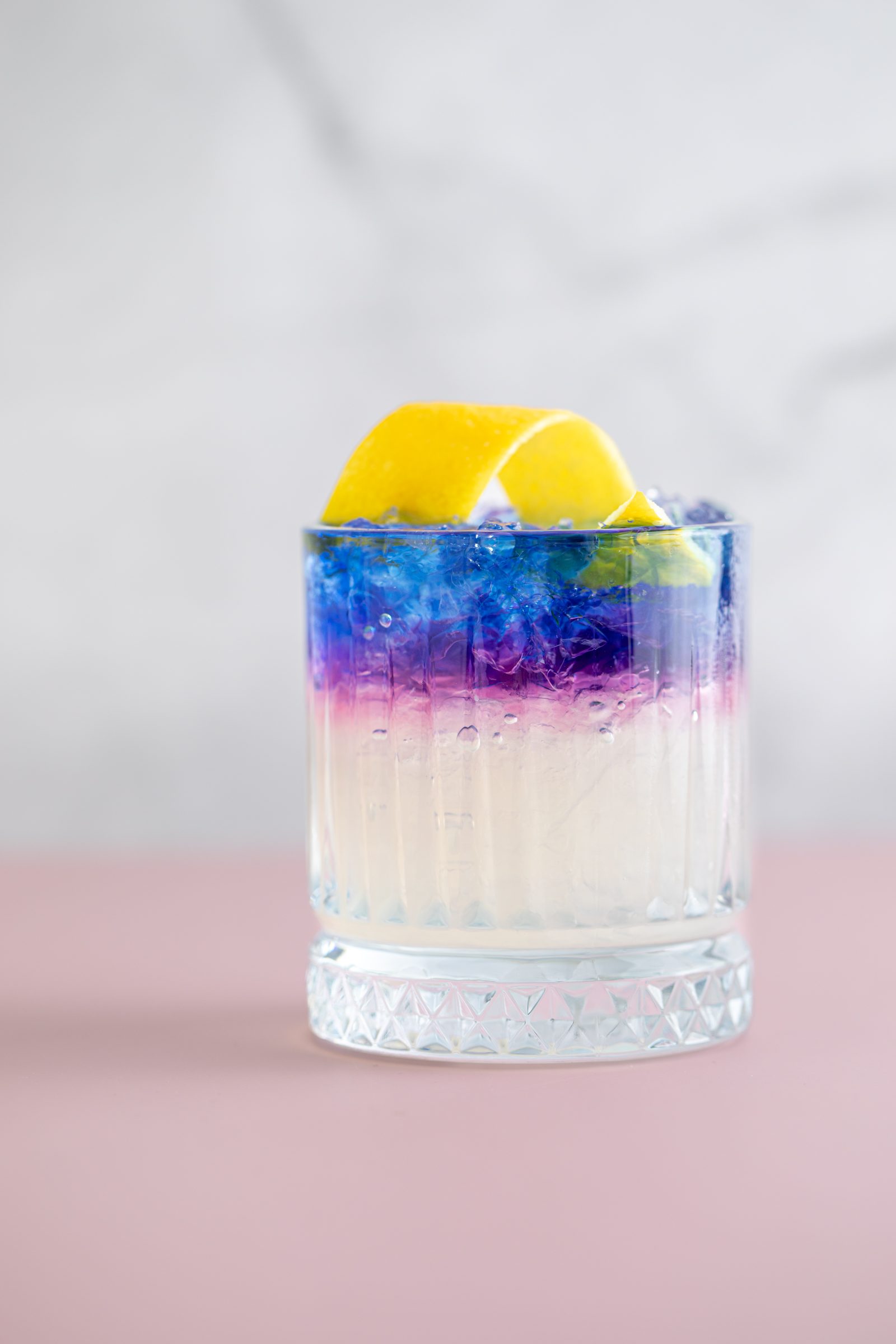 Violett und weiß geschichteter Cocktail mit Zitronenzeste im Kristallglas vor rosa und grauem Hintergrund, Cocktailfoto für das Ludwig in Mosbach