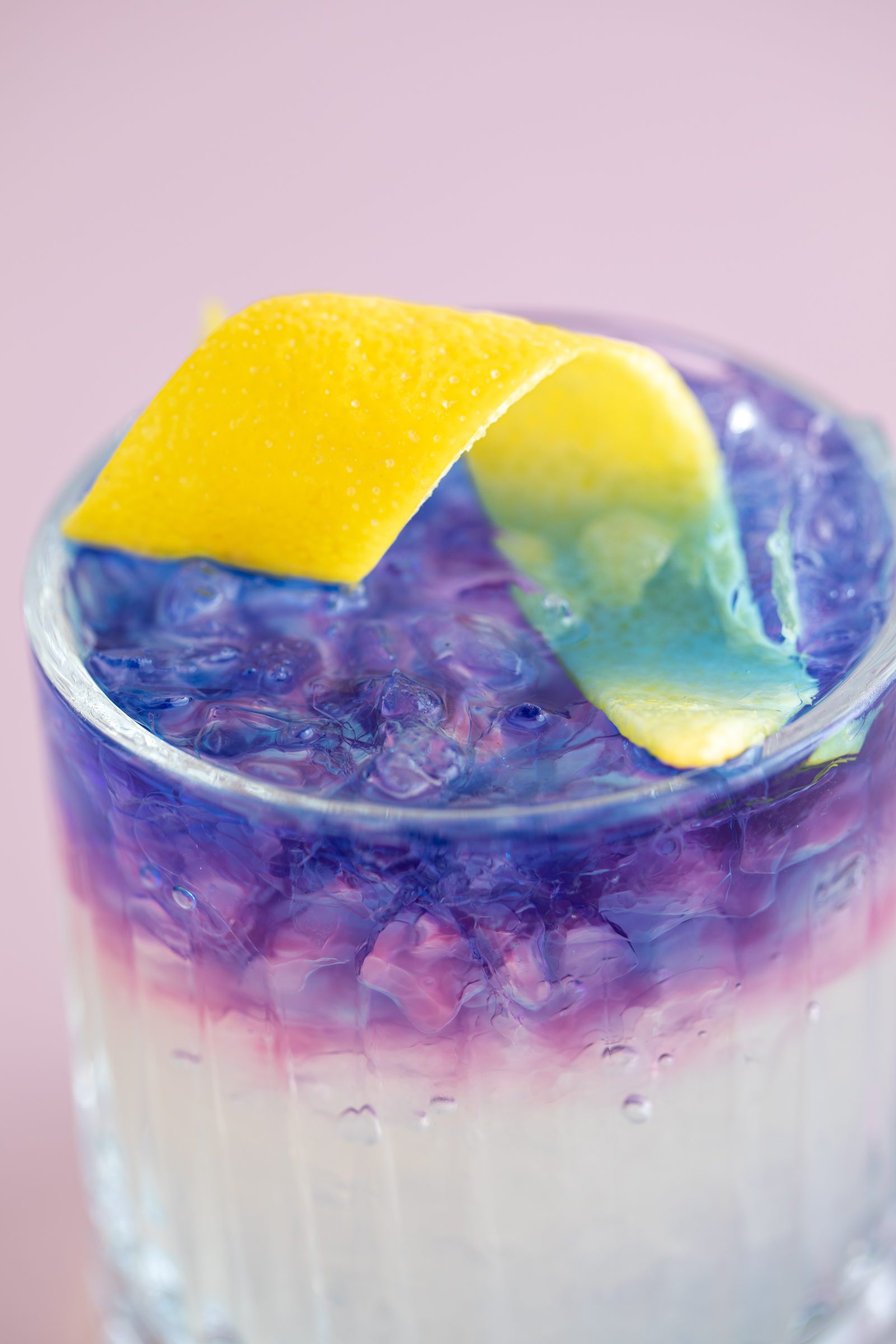 Nahaufnahme eines violett geschichteten Cocktails mit Eis und Zitronenzeste vor rosa Hintergrund, Cocktailfoto für das Ludwig in Mosbach