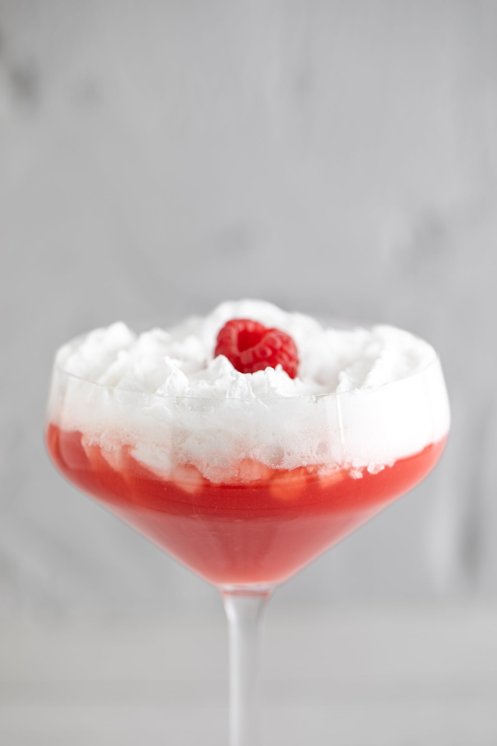 Roter Cocktail mit weißer Schaumkrone und Himbeere in einer Cocktailschale vor grauem Hintergrund, Cocktailfoto für das Ludwig in Mosbach