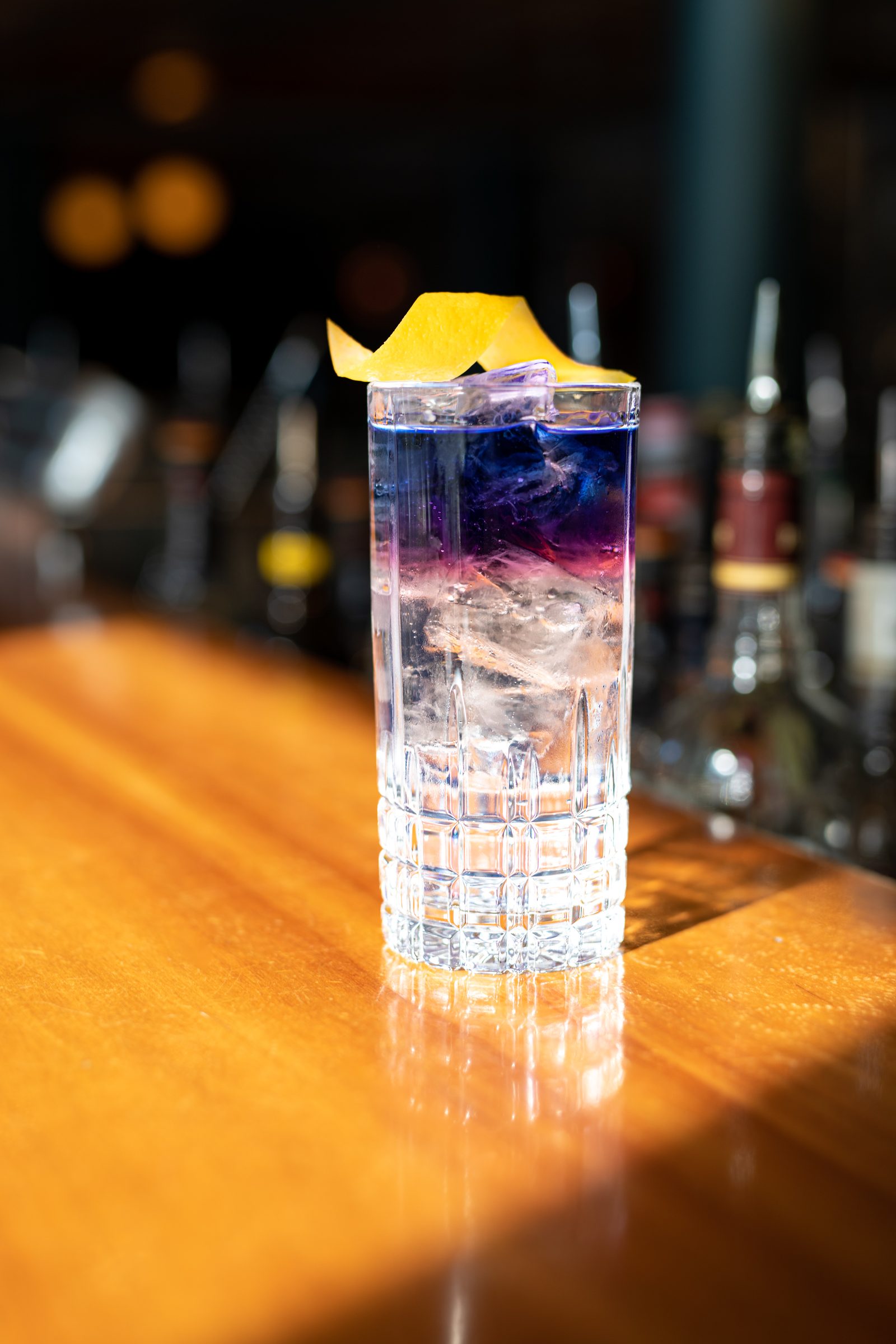 Violett geschichteter Longdrink mit Zitronenzeste im Sonnenlicht auf dem Holztresen der Bar des Ludwig in Mosbach