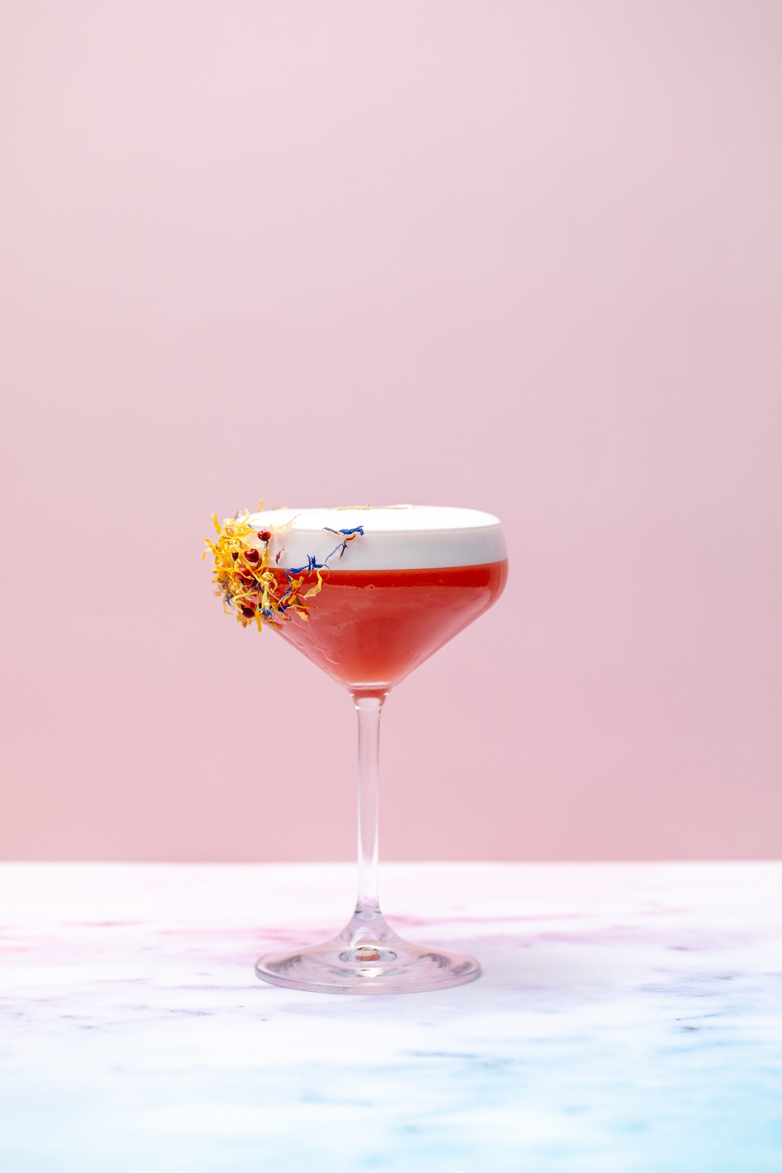 Roter Cocktail mit Schaum und Blütengarnitur in einer Cocktailschale auf Marmor vor rosa Hintergrund, Cocktailfoto für das Ludwig in Mosbach