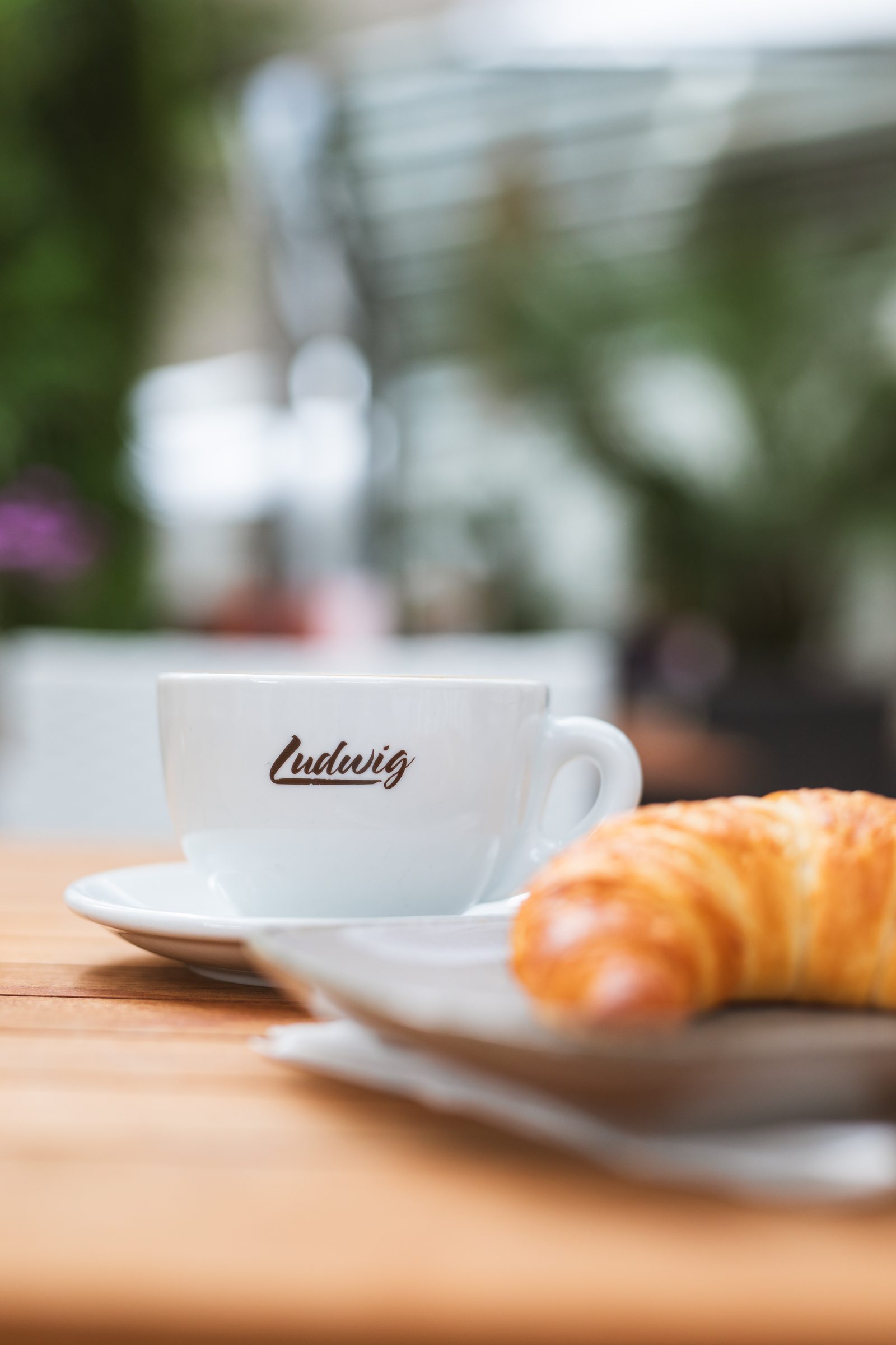 Kaffeetasse mit Ludwig-Schriftzug und Croissant auf einem Holztisch im Ludwig in Mosbach