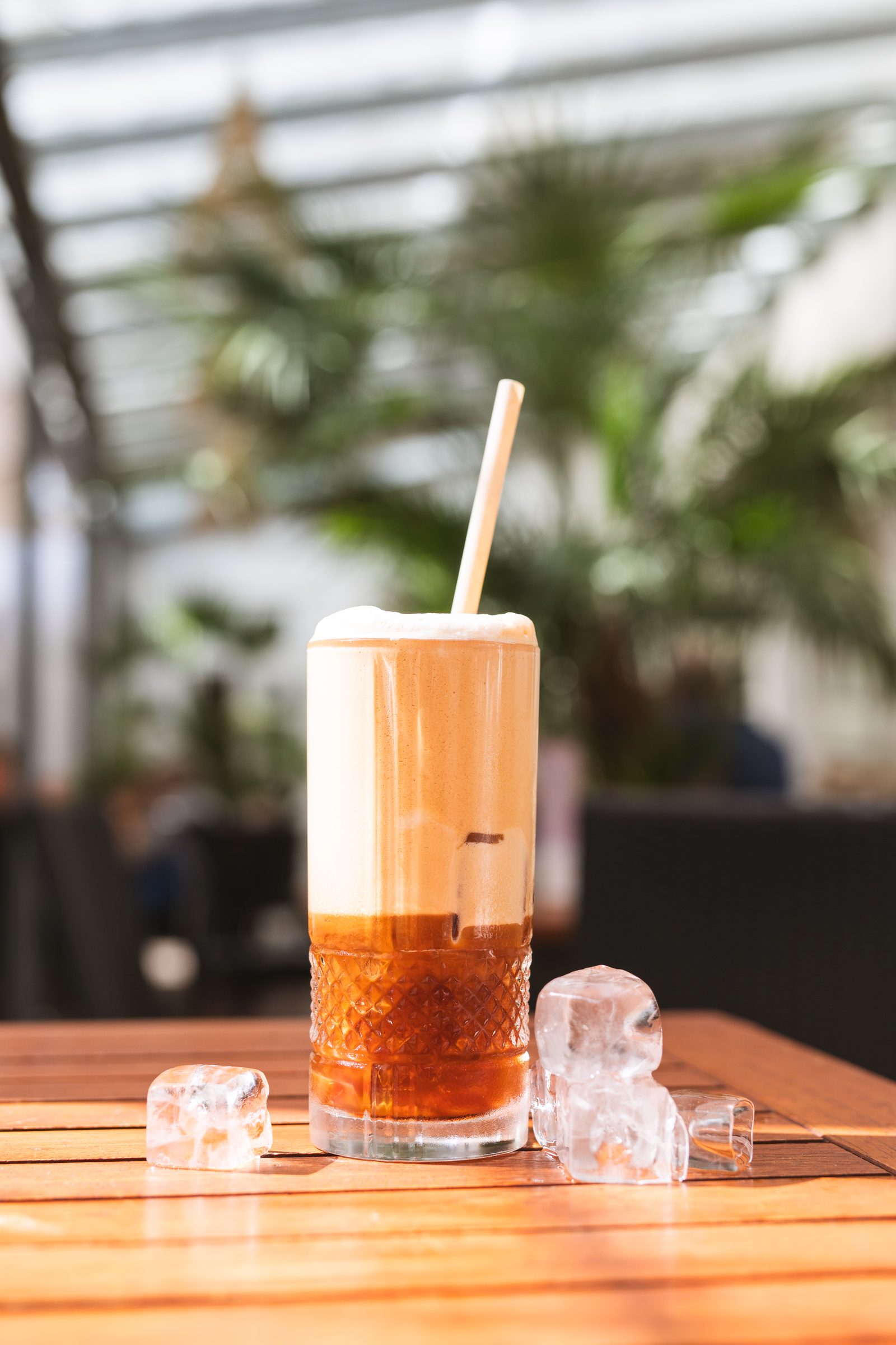 Kalter Kaffeedrink mit Schaumkrone und Papierstrohhalm im hohen Kristallglas, daneben Eiswürfel auf einem Holztisch im Ludwig in Mosbach