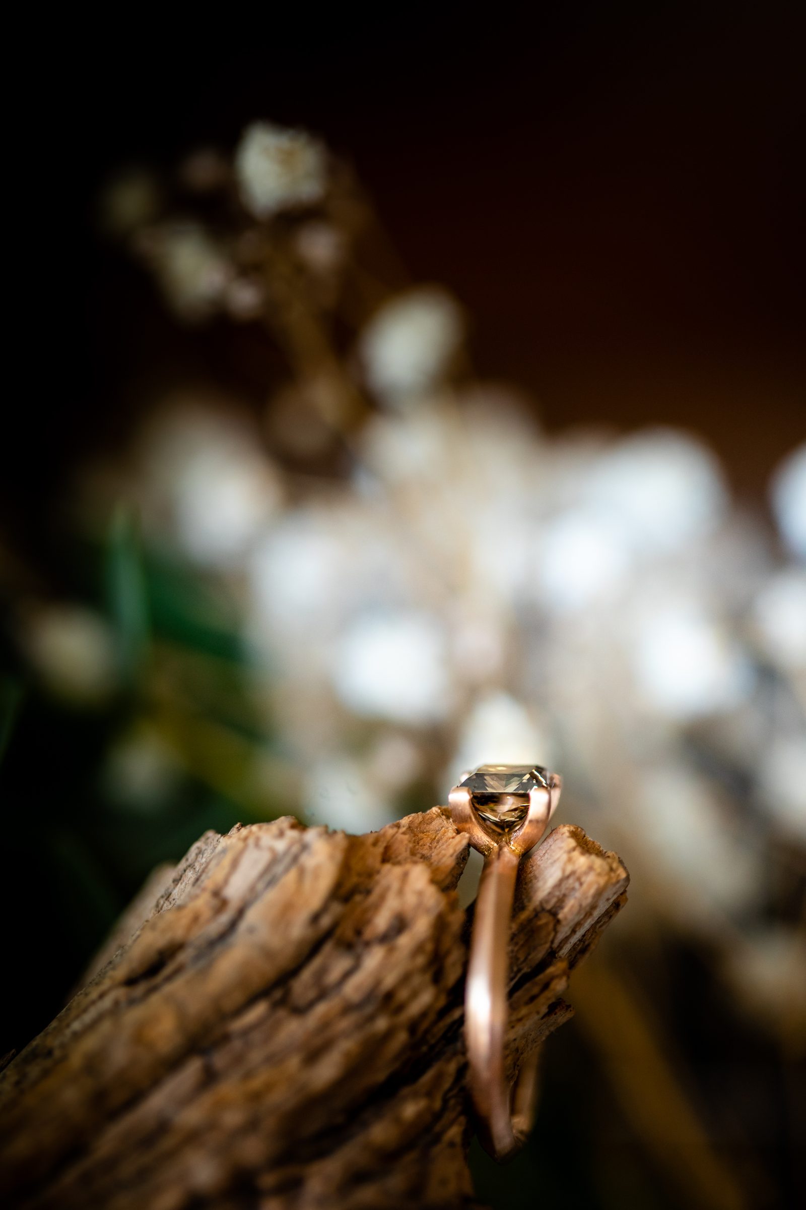 Schmaler Ring aus Roségold mit einem Diamanten an einem Stück Holz, dahinter unscharfe weiße Blüten, Schmuck von Marion Knorr