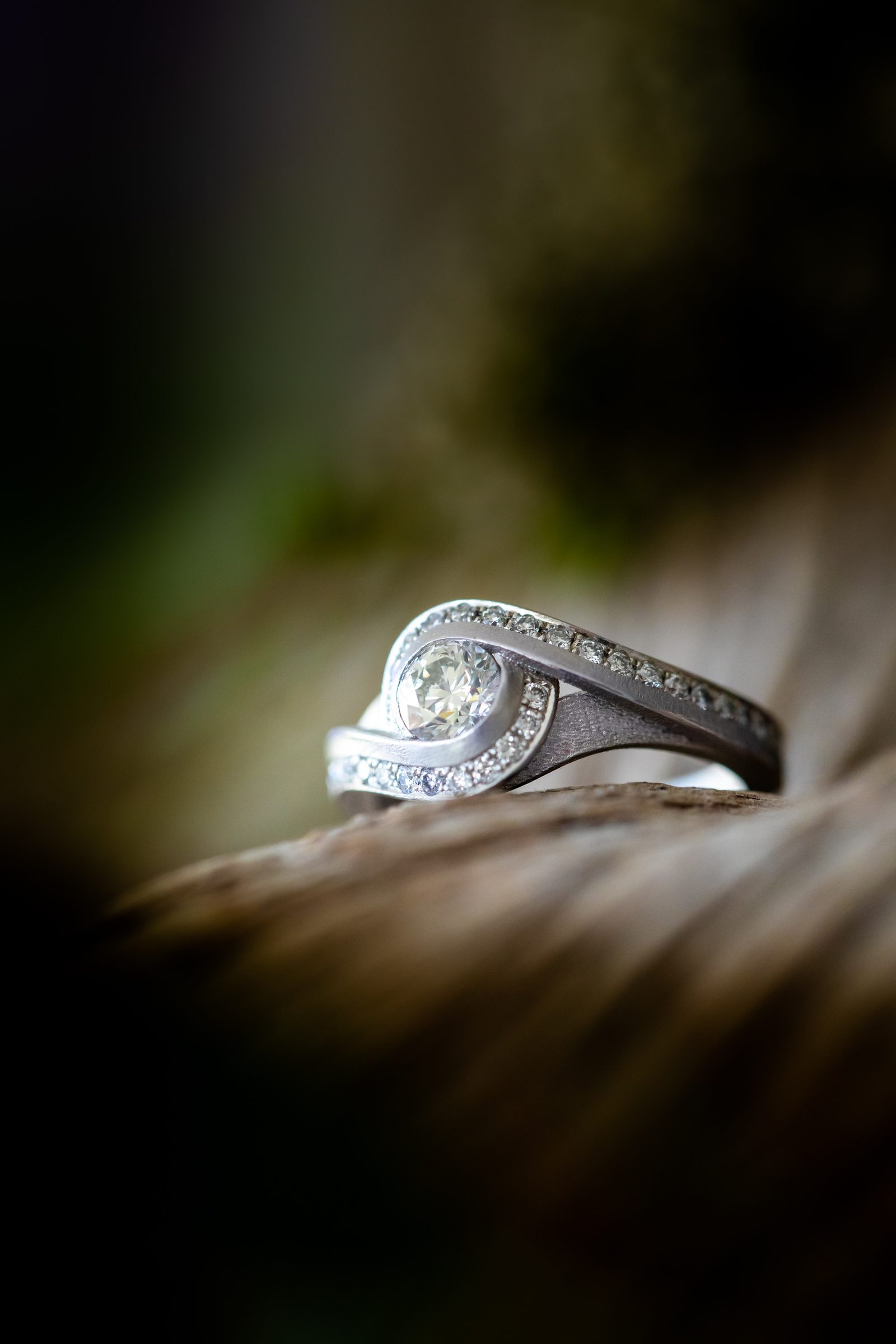 Silberfarbener Ring mit geschwungener Fassung und kleinen Diamanten auf einem Stück Holz vor dunkelgrünem Hintergrund, Schmuck von Marion Knorr