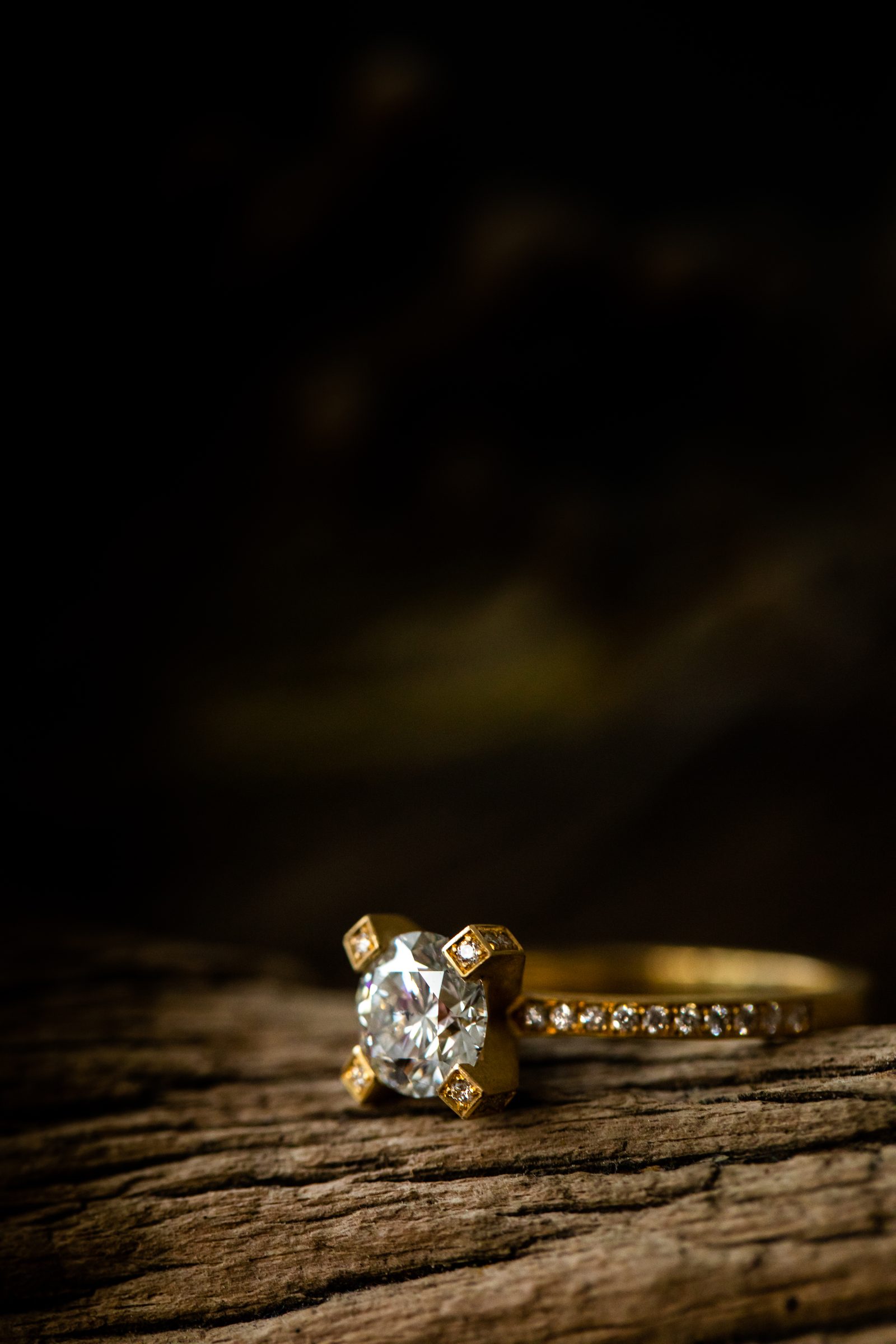 Goldring mit Diamant in Krappenfassung und kleinen Steinen auf der Ringschiene auf einem Stück Holz vor dunklem Hintergrund, Schmuck von Marion Knorr