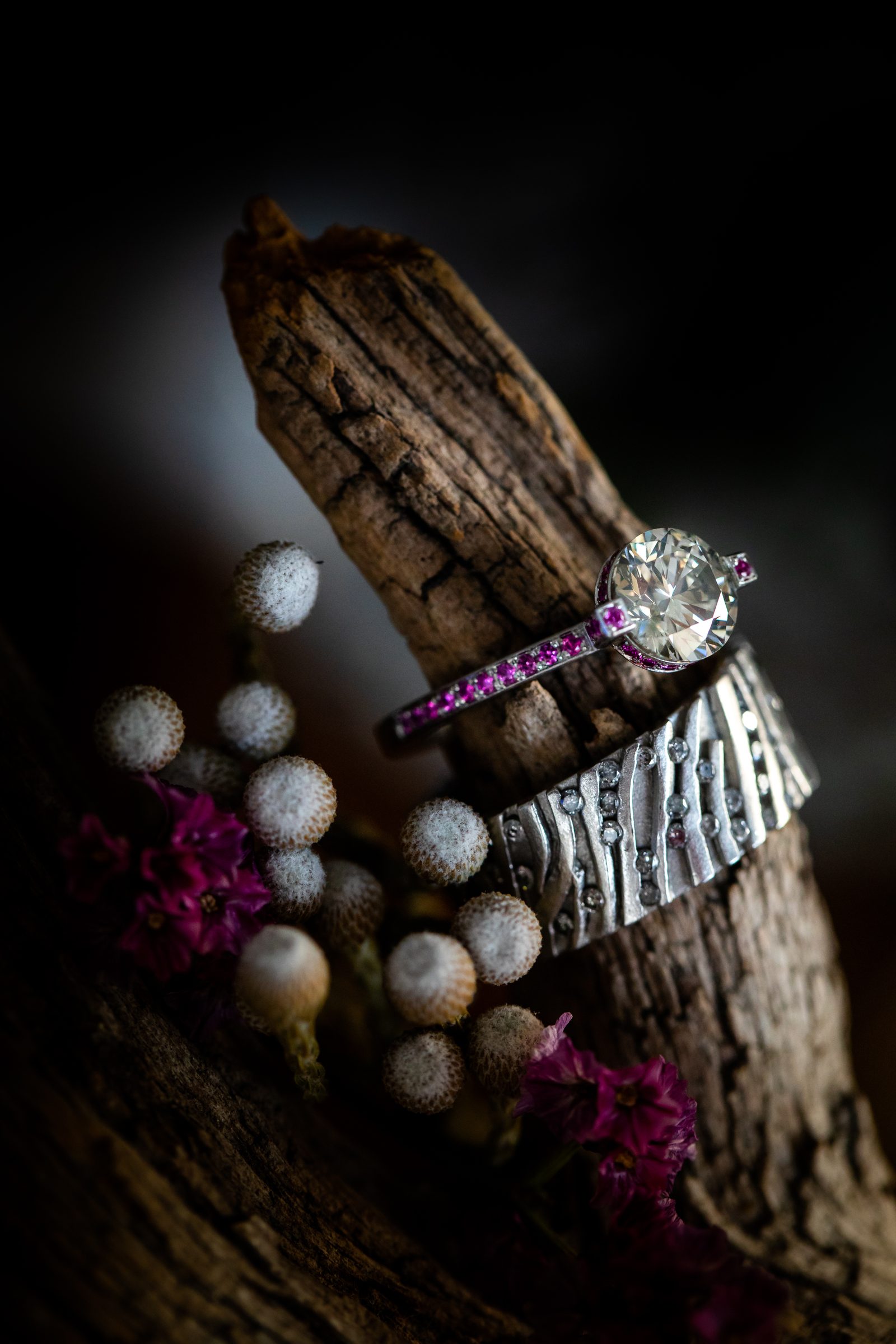 Ring mit Diamant und pinkfarbenen Steinen über einem breiten strukturierten Ring an Treibholz, davor Trockenblumen in Weiß und Pink, Schmuck von Marion Knorr