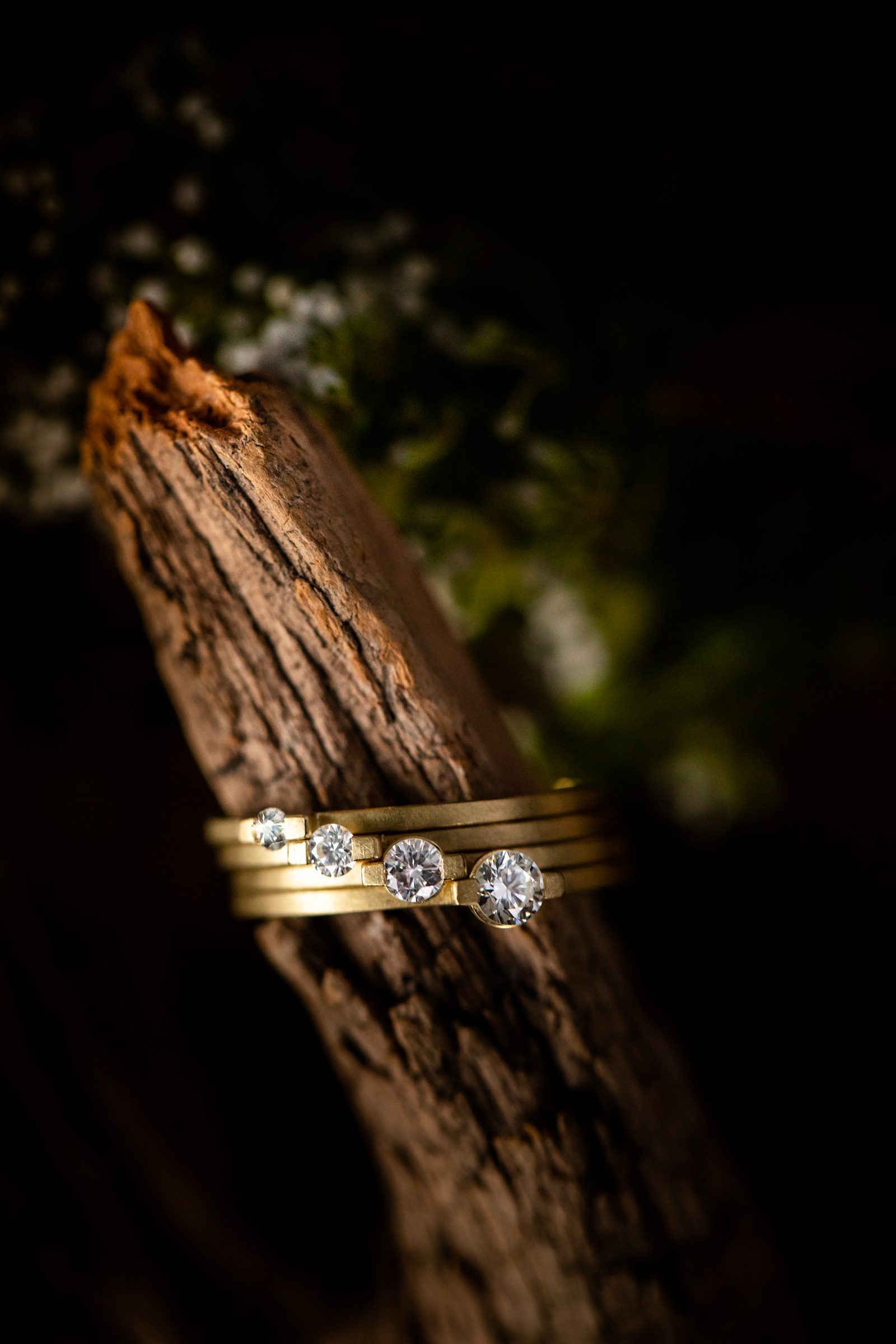 Mehrere schmale Goldringe mit Diamanten übereinander an einem Stück Holz, dahinter Grün und kleine weiße Blüten, Schmuck von Marion Knorr