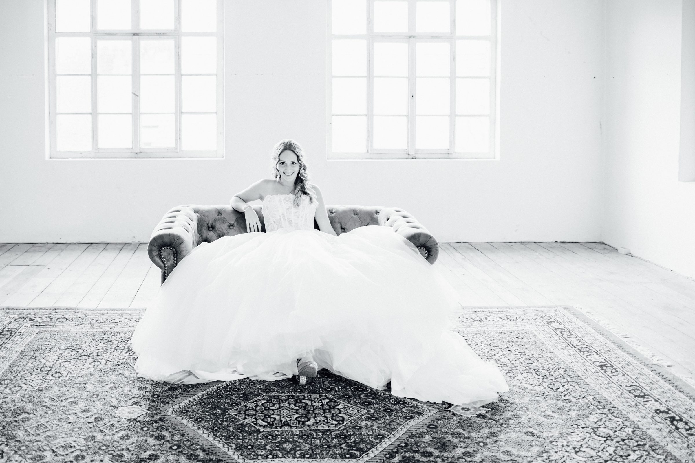 Schwarzweißfoto einer Frau in einem Brautkleid mit weitem Tüllrock auf einem Chesterfield-Sofa in einem hellen Loft mit Sprossenfenstern, Kollektionsfoto für MarryMe Brautmode