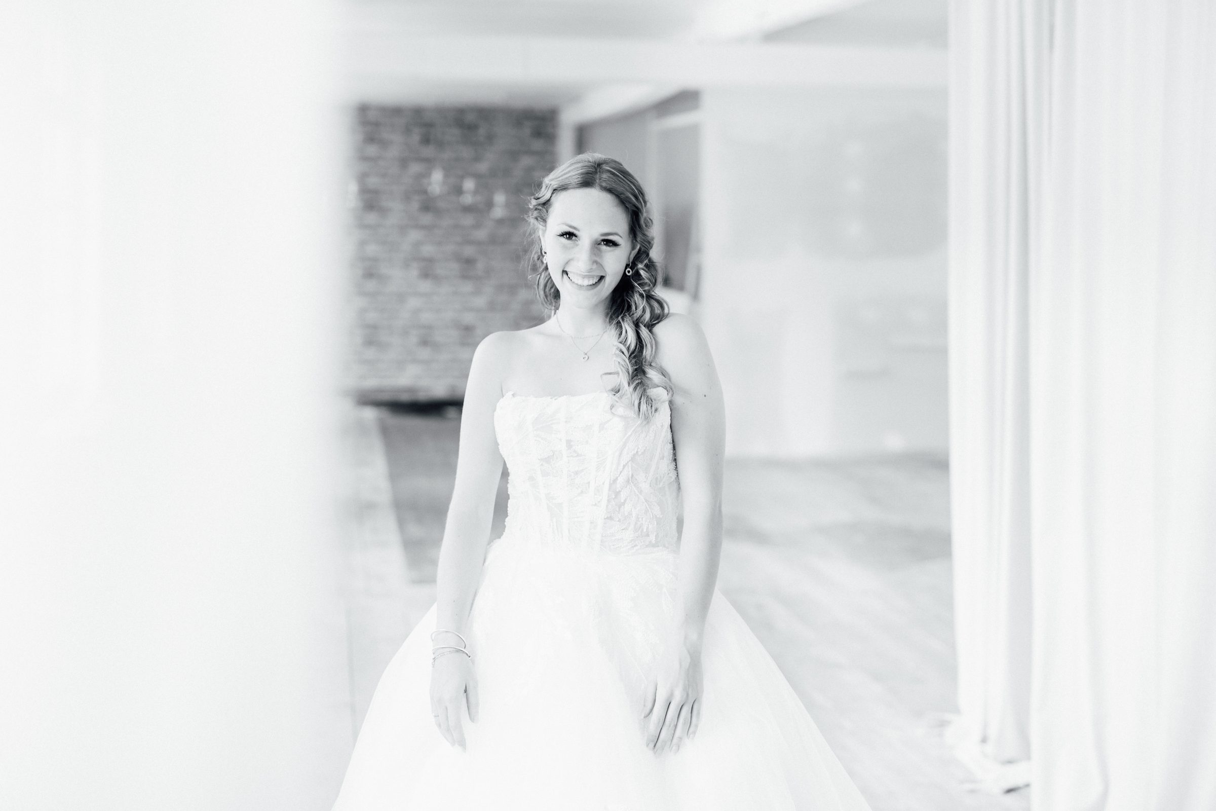 Schwarzweißfoto einer lächelnden Frau in einem trägerlosen Brautkleid mit Spitzencorsage zwischen hellen Vorhängen, Kollektionsfoto für MarryMe Brautmode