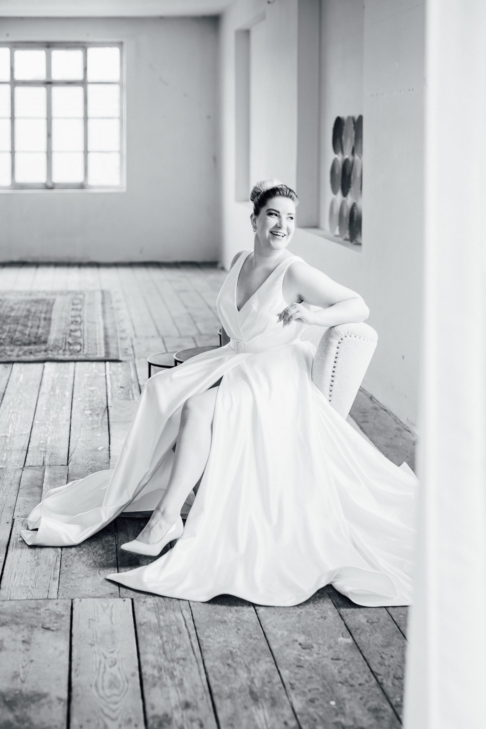 Schwarzweißfoto einer Frau in einem Brautkleid mit V-Ausschnitt und Beinschlitz auf einem Sessel am Fenster eines Lofts, Kollektionsfoto für MarryMe Brautmode