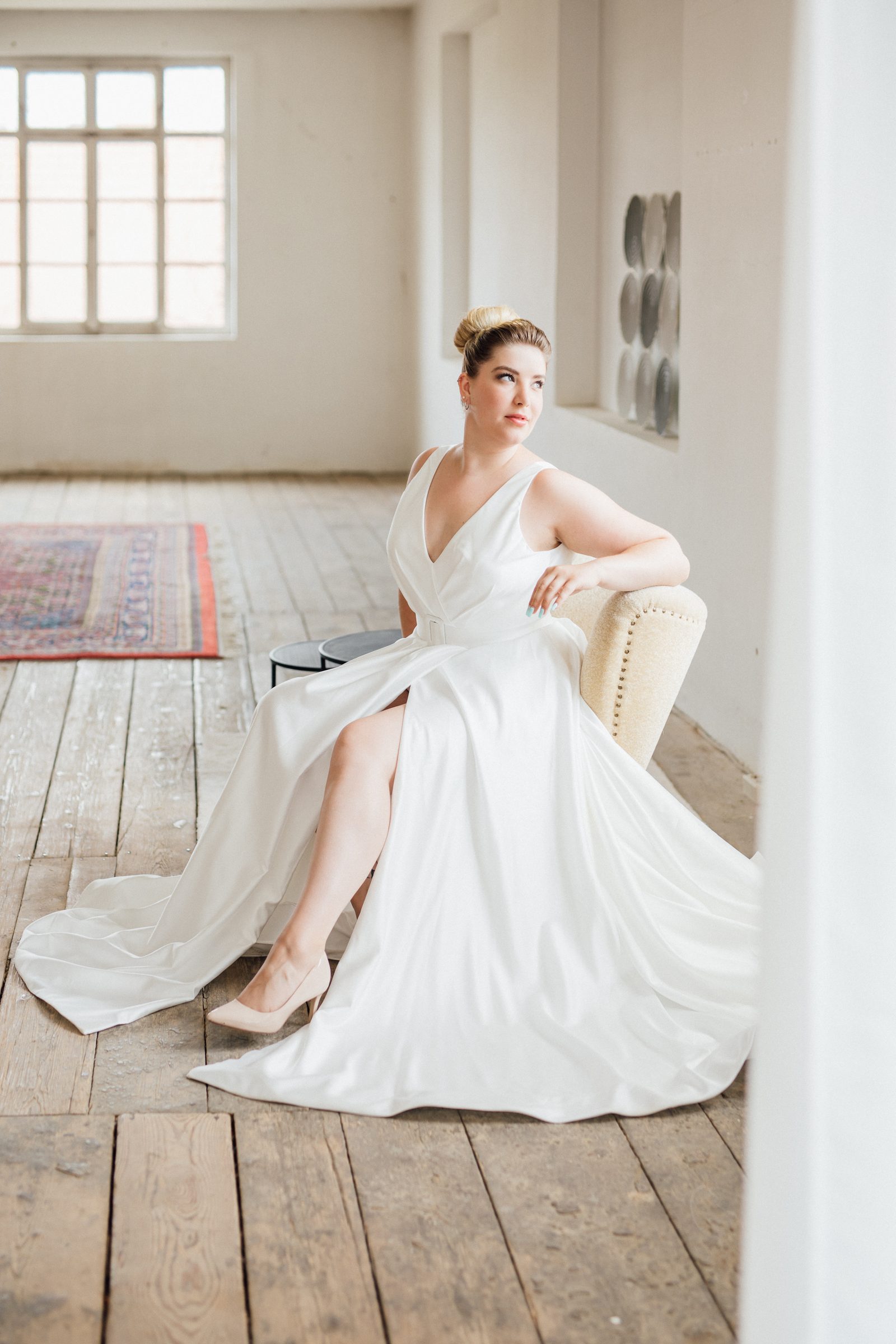 Frau in einem schlichten Satin-Brautkleid mit V-Ausschnitt und Beinschlitz sitzt auf einem Sessel in einem hellen Loft mit Holzdielen, Kollektionsfoto für MarryMe Brautmode