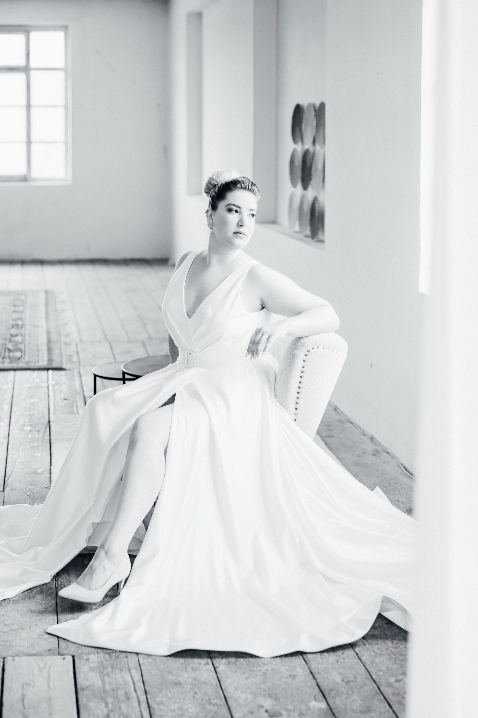 Schwarzweißfoto einer Frau, die in einem Brautkleid mit V-Ausschnitt und weit ausgebreitetem Rock auf einem Sessel sitzt und zur Seite blickt, Kollektionsfoto für MarryMe Brautmode