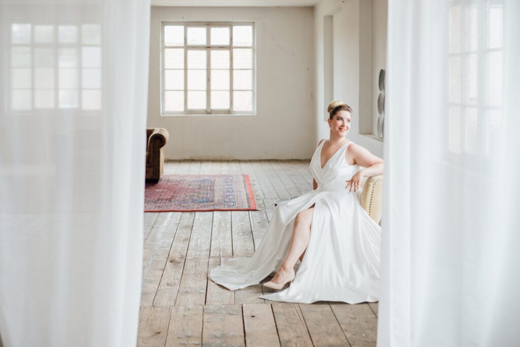 Frau in einem Satin-Brautkleid mit V-Ausschnitt sitzt auf einem Sessel in einem hellen Loft, eingerahmt von weißen Vorhängen, Kollektionsfoto für MarryMe Brautmode