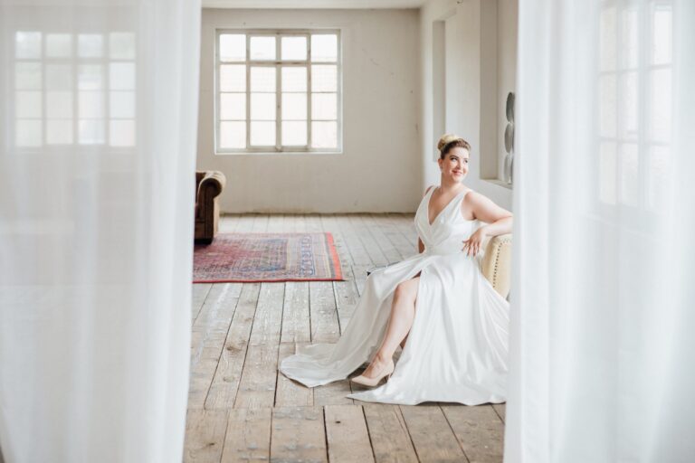Frau in einem Satin-Brautkleid mit V-Ausschnitt sitzt auf einem Sessel in einem hellen Loft, eingerahmt von weißen Vorhängen, Kollektionsfoto für MarryMe Brautmode