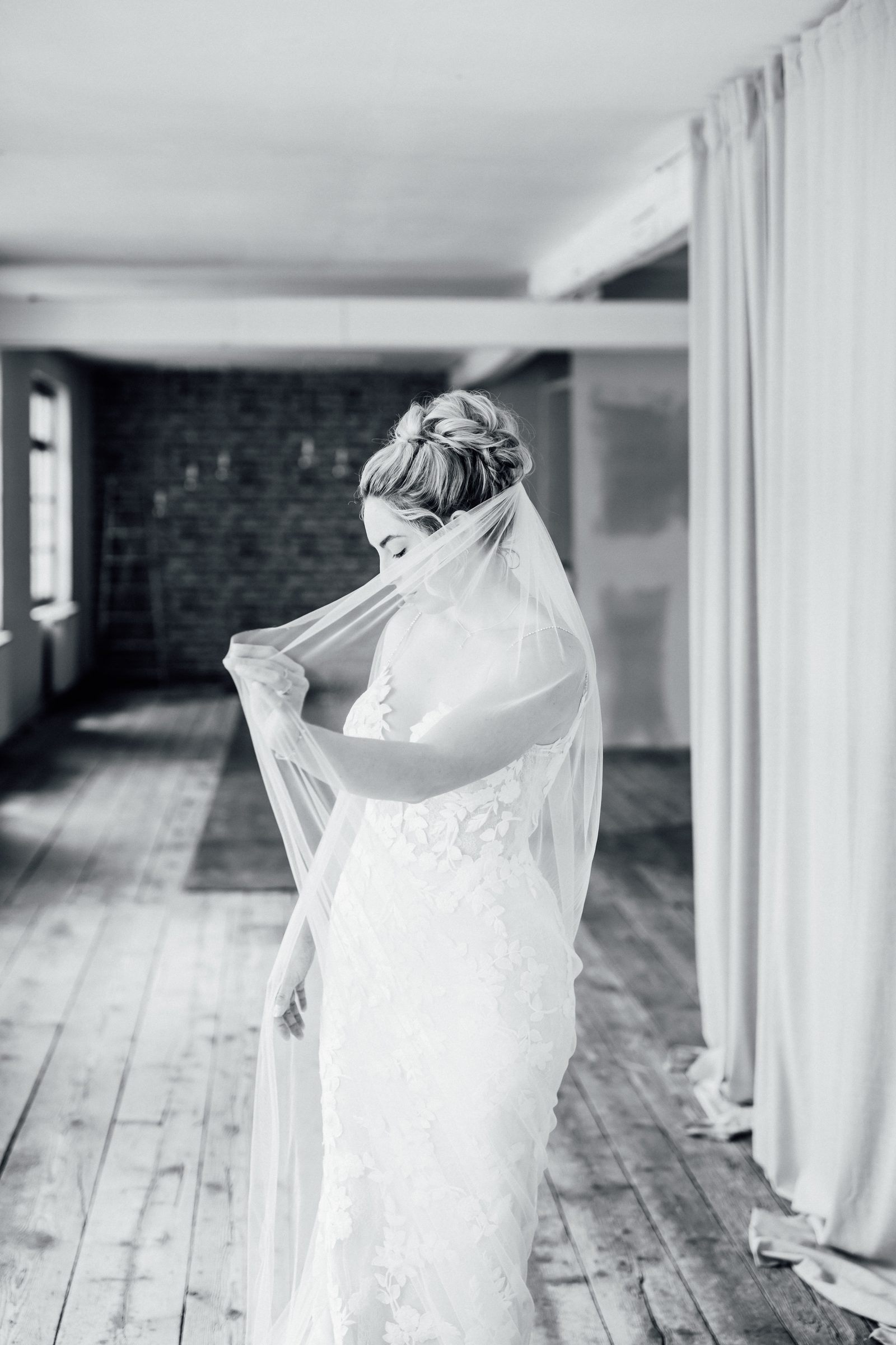 Schwarzweißfoto einer Frau in einem Spitzenbrautkleid, die ihren langen Schleier vor das Gesicht hebt, in einem Loft mit Holzdielen und Vorhängen, Kollektionsfoto für MarryMe Brautmode