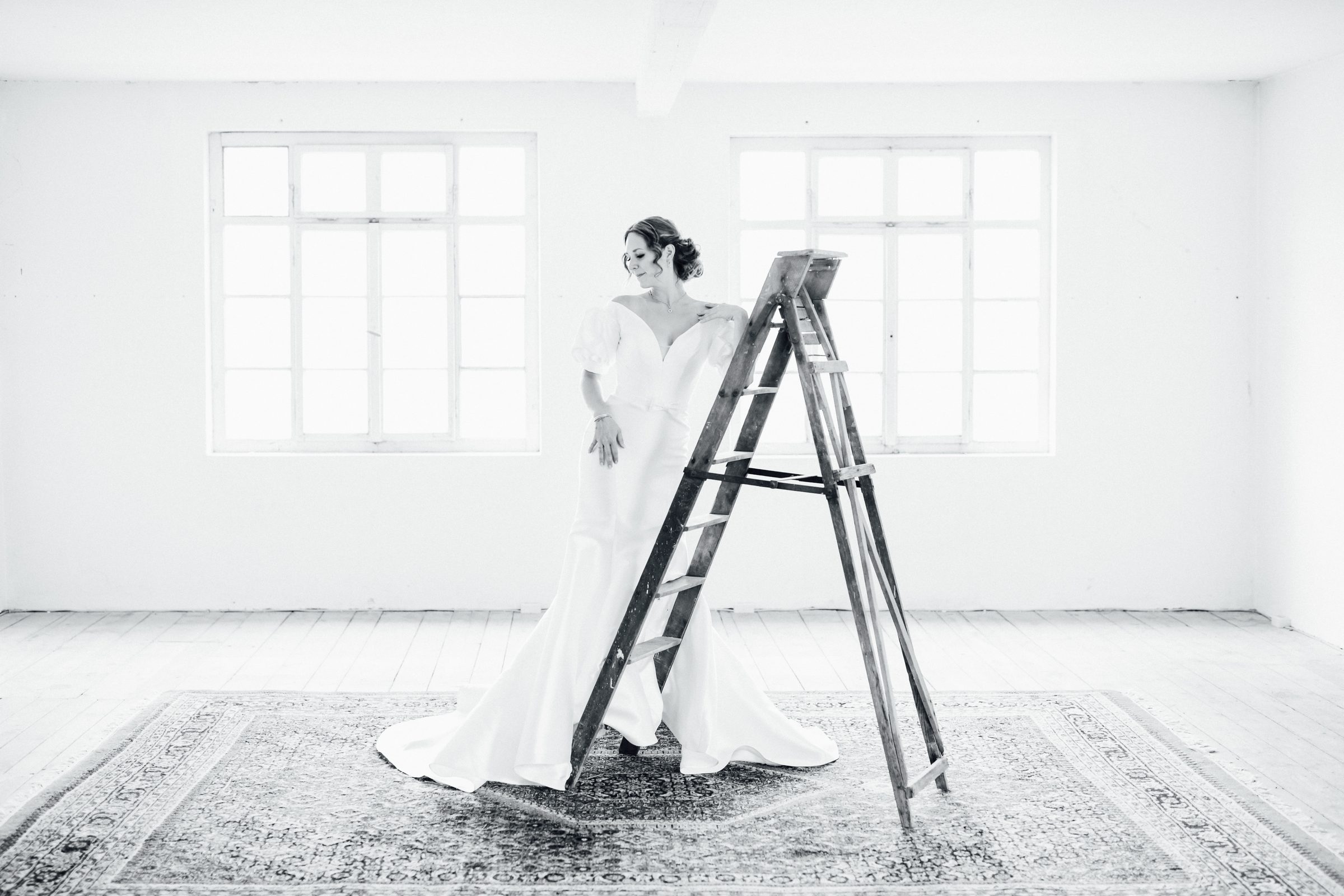 Schwarzweißfoto einer Frau in einem Brautkleid mit Puffärmeln und Schleppe neben einer alten Holzleiter auf einem Teppich vor Sprossenfenstern, Kollektionsfoto für MarryMe Brautmode