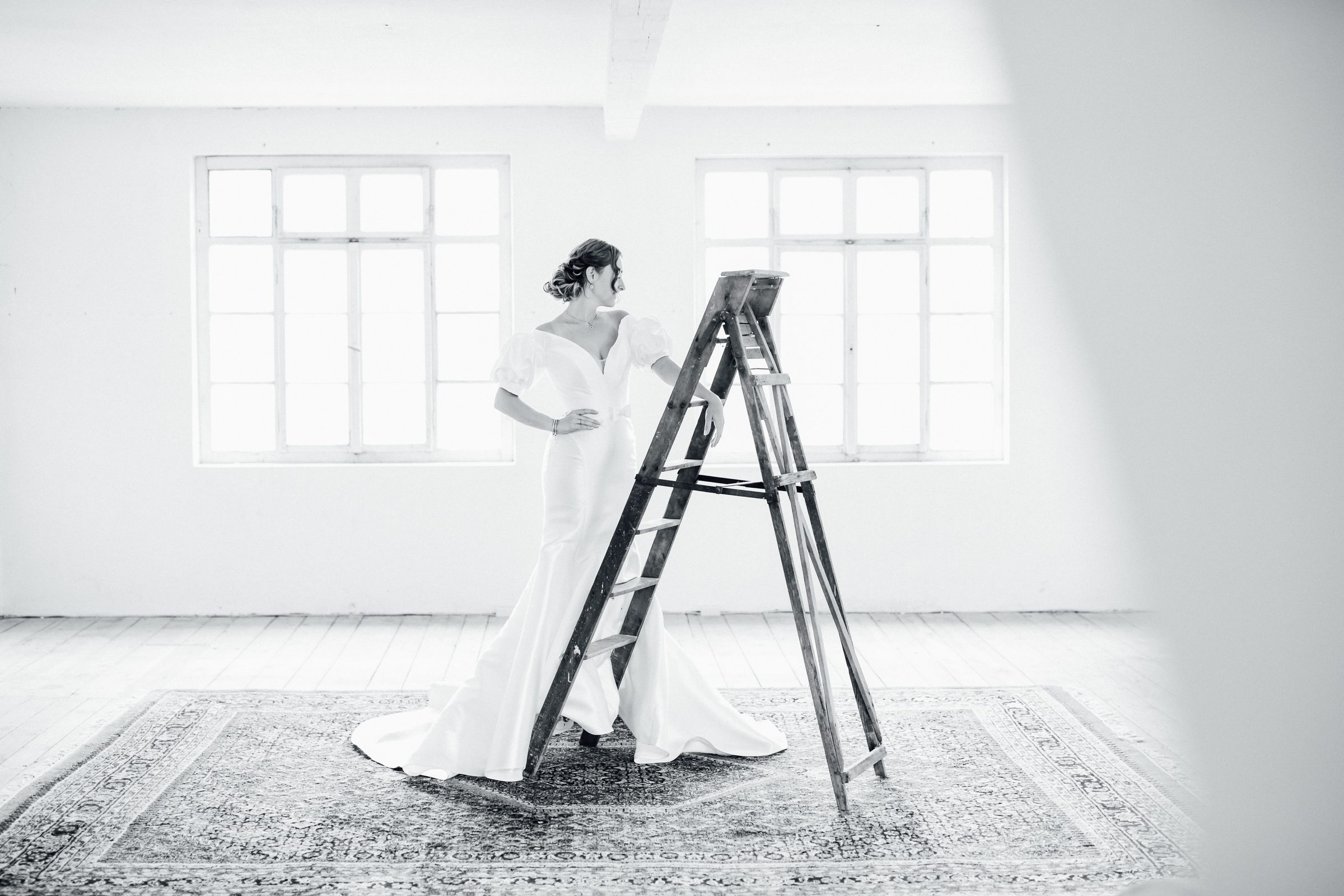 Schwarzweißfoto einer Frau in einem Brautkleid mit Puffärmeln, die eine Hand in die Hüfte stützt und sich an eine Holzleiter lehnt, heller Raum mit Sprossenfenstern, Kollektionsfoto für MarryMe Brautmode