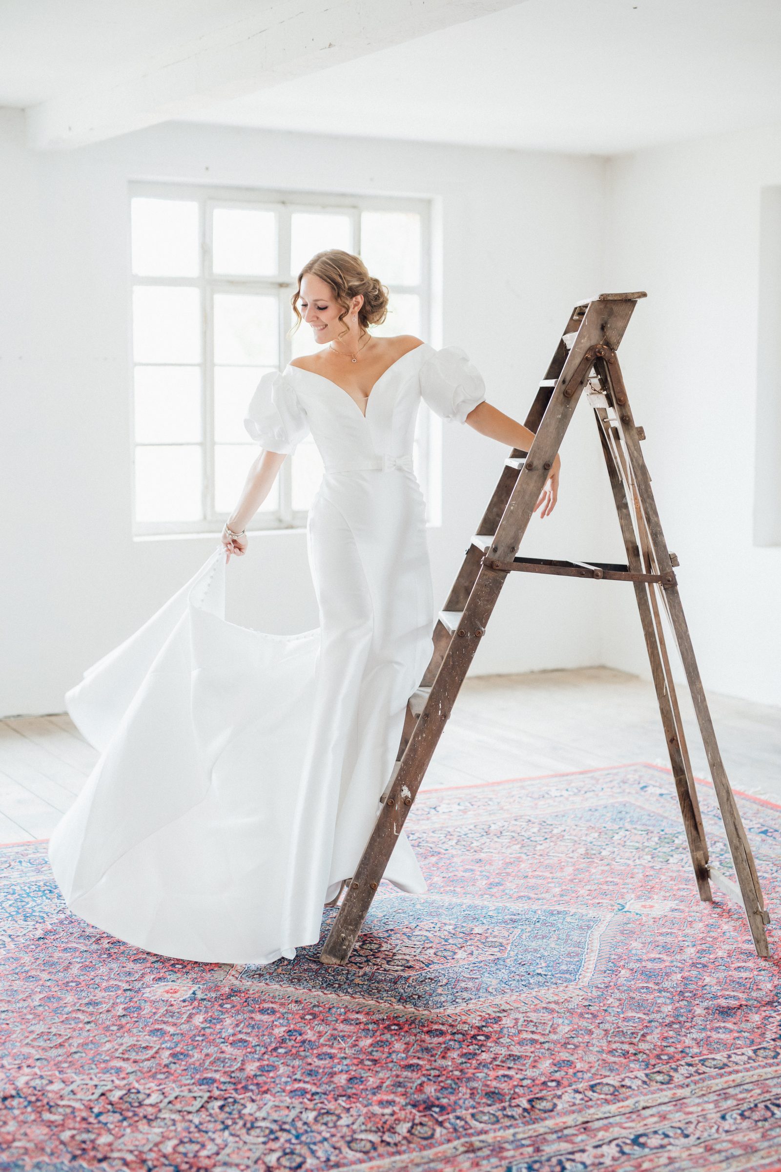 Frau in einem Brautkleid mit Puffärmeln hält den Rock und lehnt an einer Holzleiter auf einem roten Orientteppich in einem hellen Loft, Kollektionsfoto für MarryMe Brautmode