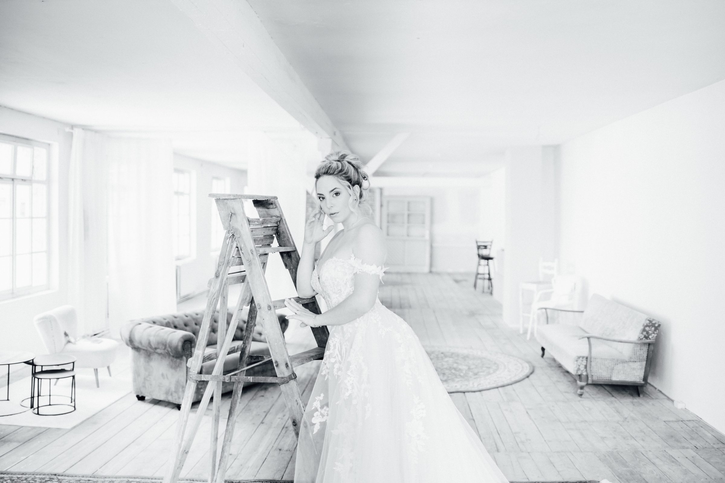 Schwarzweißfoto einer Frau in einem schulterfreien Spitzenbrautkleid an einer Holzleiter in einem Loft mit Sofas und Sesseln, Kollektionsfoto für MarryMe Brautmode