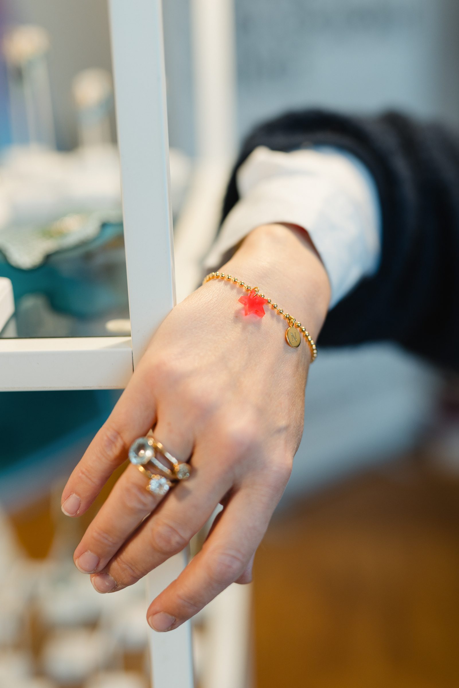 Hand mit zartem Goldarmband mit rotem Stern und zwei Ringen an einem weißen Regal, Detailaufnahme für Neue Zeiten