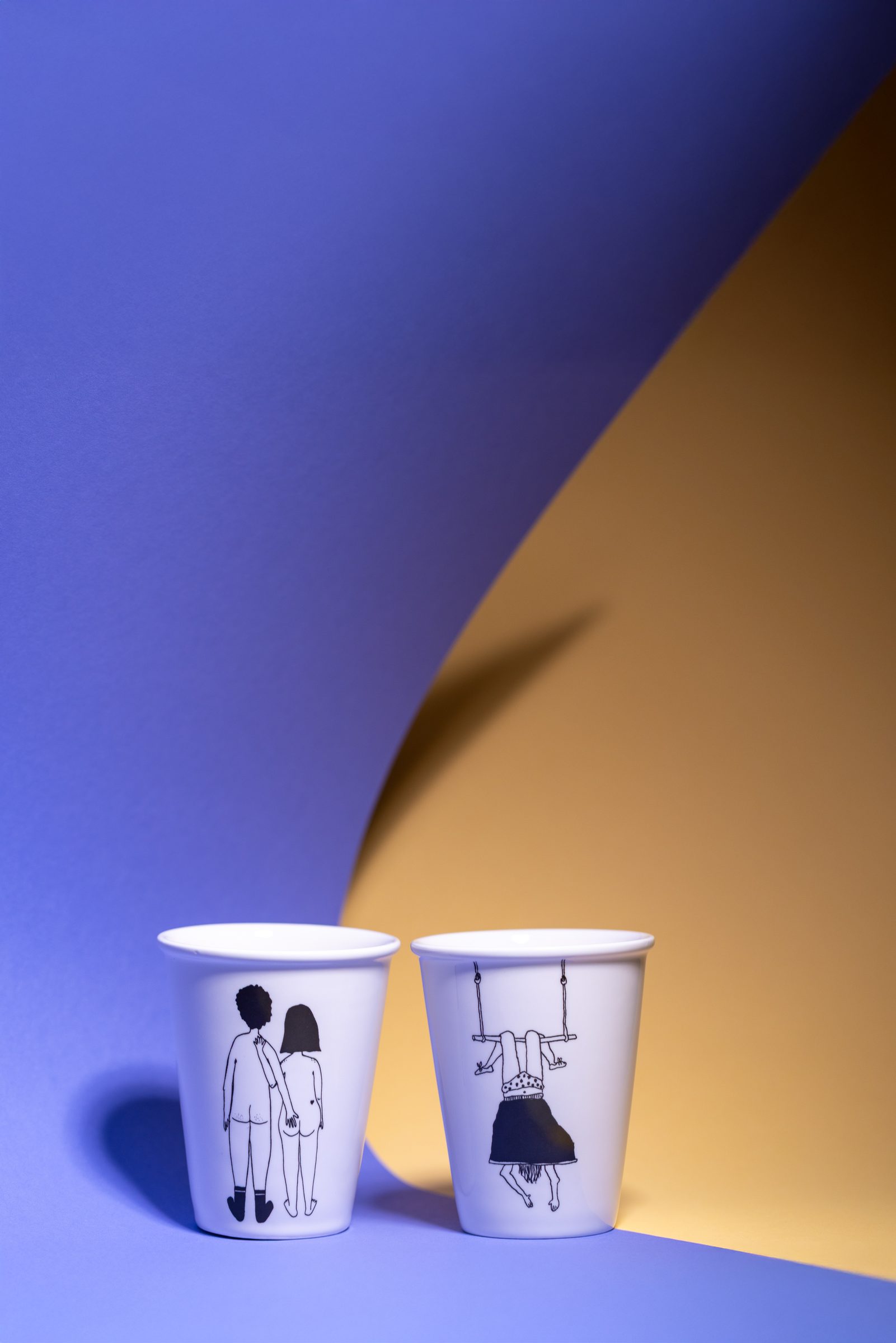 Zwei weiße Porzellanbecher mit schwarzen Zeichnungen, ein Paar und eine Figur auf einer Schaukel, vor blauem und ockerfarbenem Hintergrund, Produktfoto für Neue Zeiten