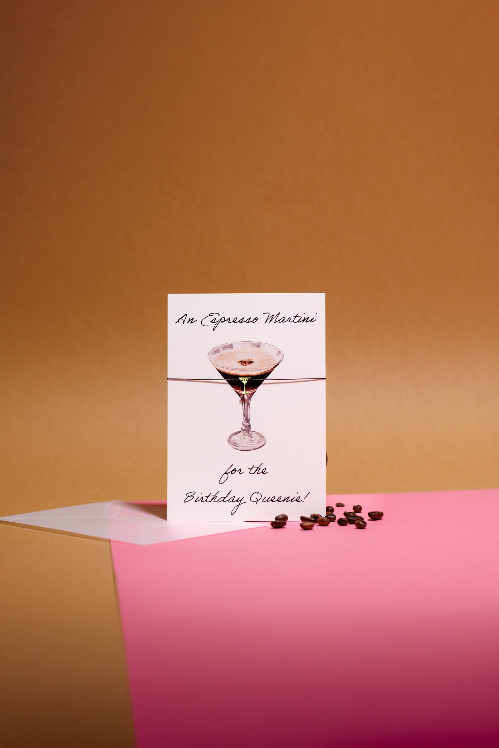 Geburtstagskarte mit gezeichnetem Cocktailglas auf rosa und ockerfarbenem Papier, daneben ein Umschlag und einige Kaffeebohnen, Produktfoto für Neue Zeiten