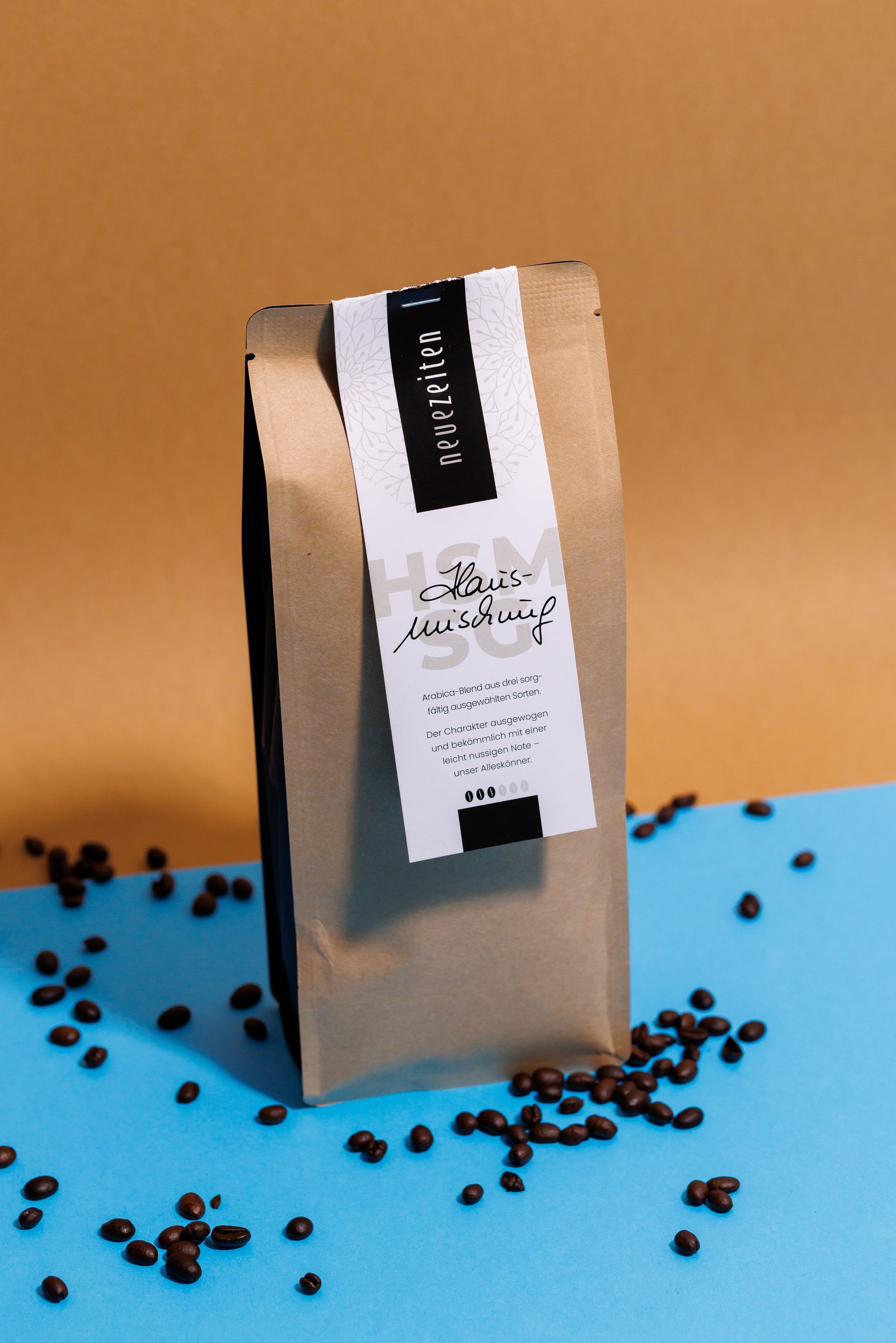 Nahaufnahme einer Kaffeepackung der Hausmischung aus der eigenen Rösterei von Neue Zeiten mit verstreuten Kaffeebohnen vor ockerfarbenem und hellblauem Hintergrund