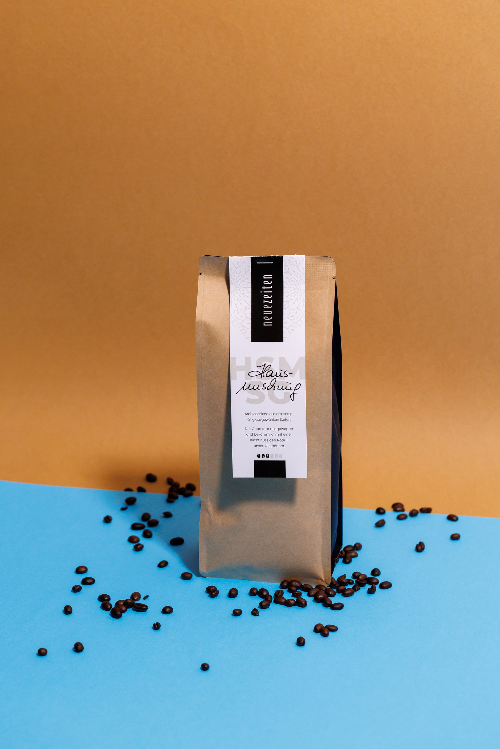 Kaffeepackung der Hausmischung aus der eigenen Rösterei von Neue Zeiten zwischen weit verstreuten Kaffeebohnen vor ockerfarbenem und hellblauem Hintergrund