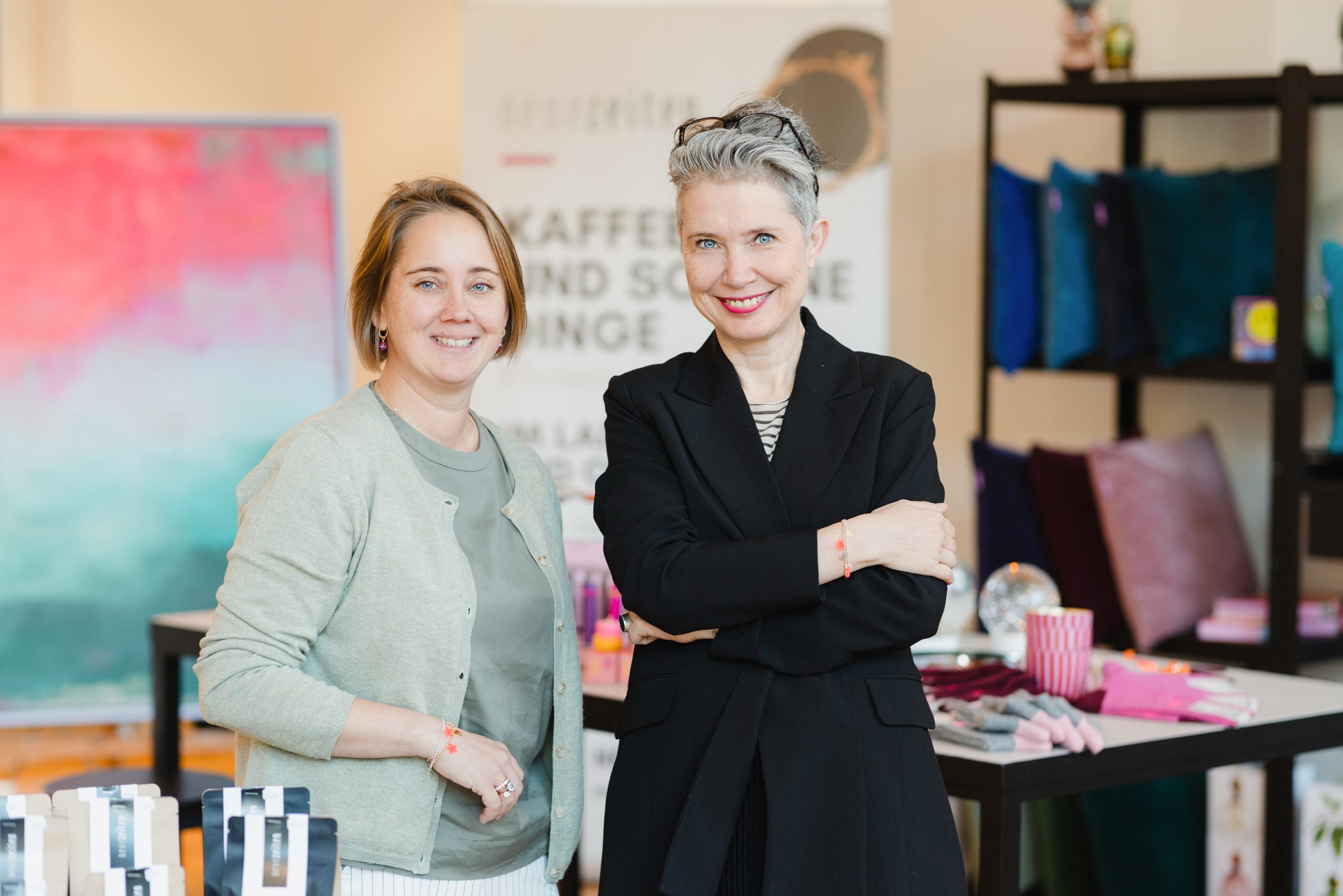 Zwei lächelnde Frauen von Neue Zeiten vor Regalen mit Kissen und Wohnaccessoires, dahinter ein Banner mit dem Schriftzug neuezeiten