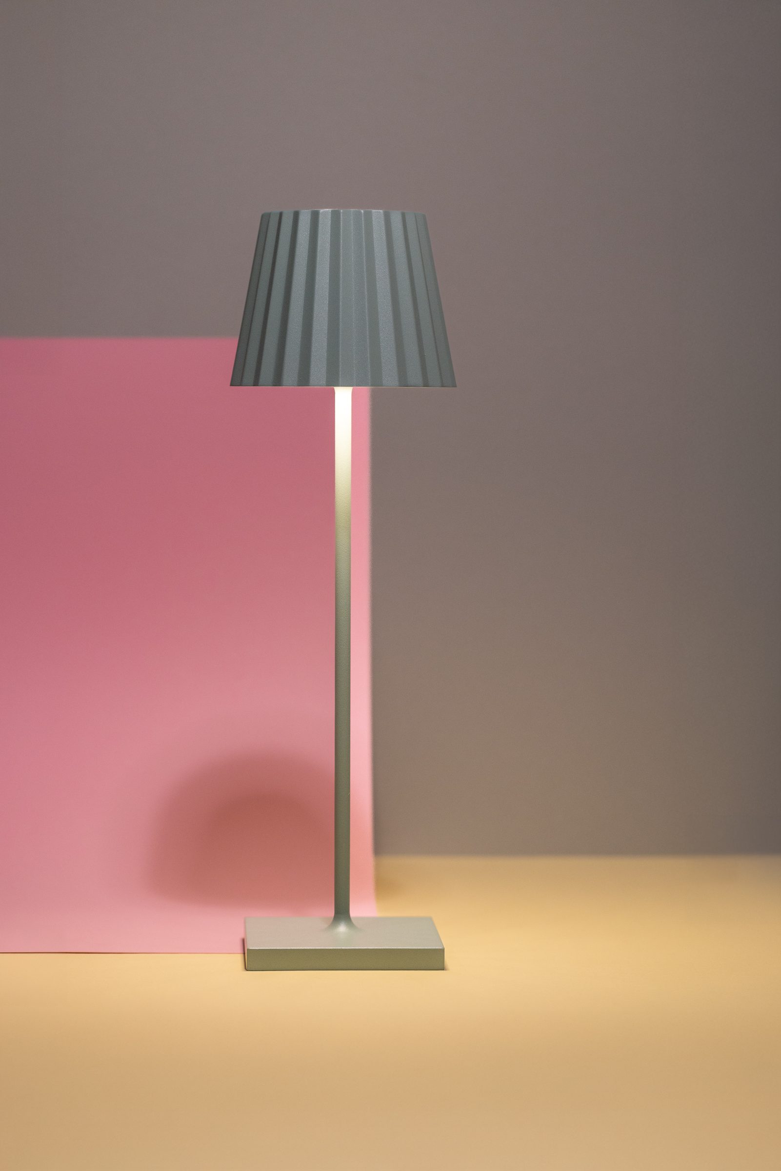Schlanke Tischleuchte mit grauem Faltenschirm vor rosa, grauem und beigem Hintergrund, Produktfoto für Neue Zeiten