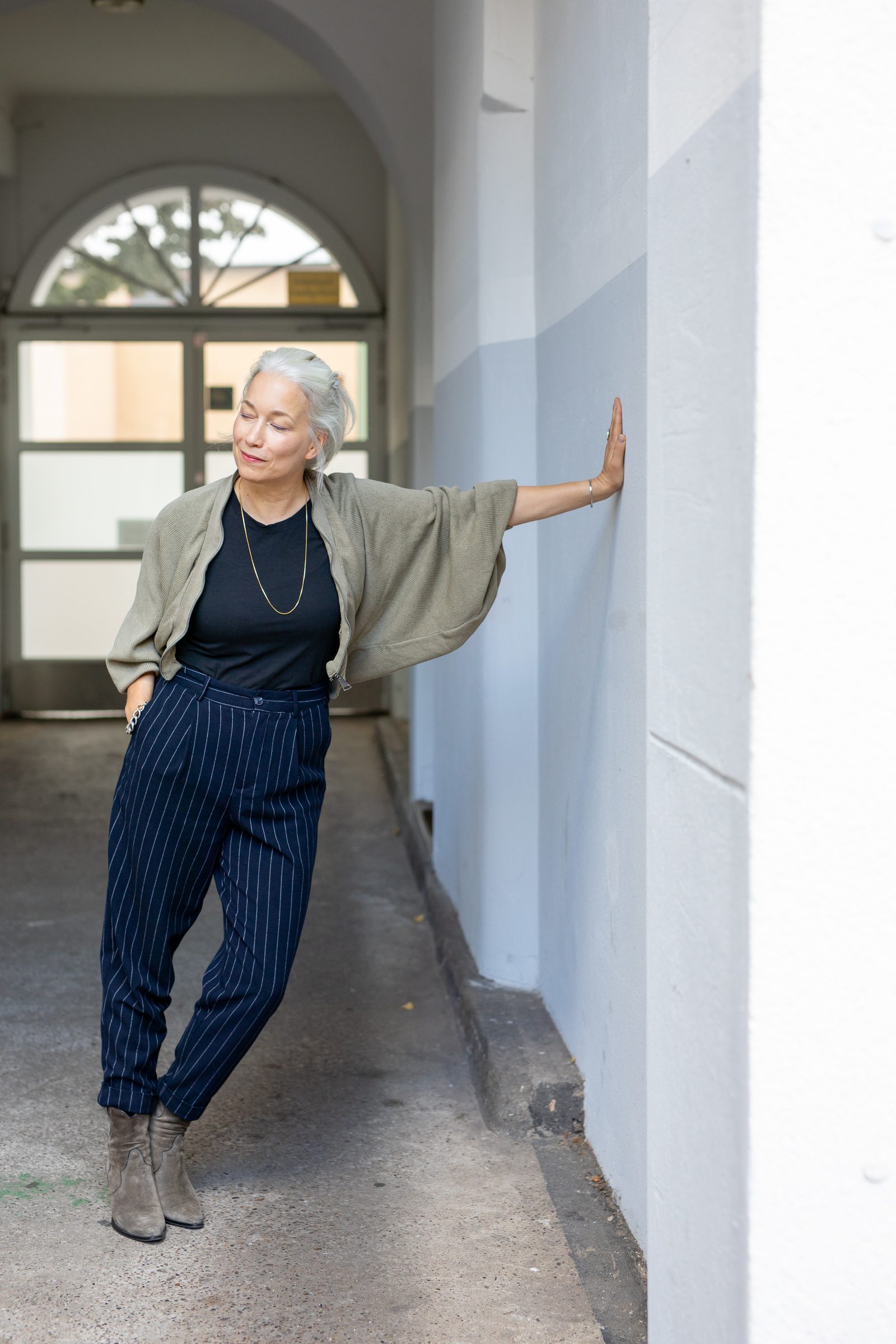 Nina Goldhammer in olivgrüner Strickjacke stützt sich in einem hellen Durchgang mit Rundbogenfenster an der Wand ab