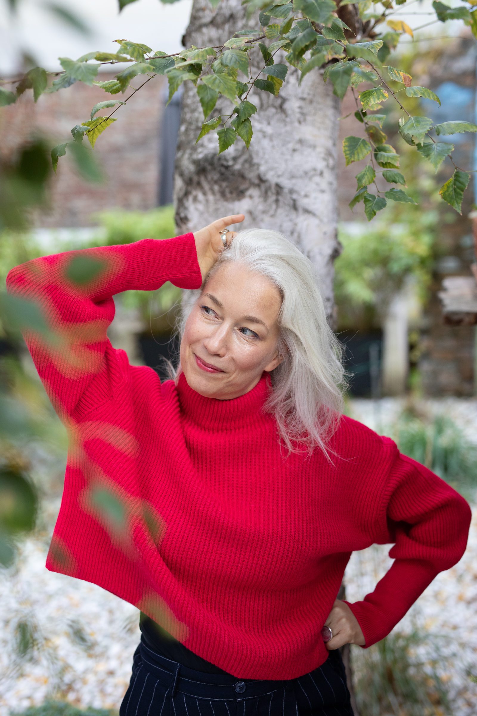 Nina Goldhammer in rotem Pullover steht unter einem Baum im Garten und legt eine Hand an den Kopf