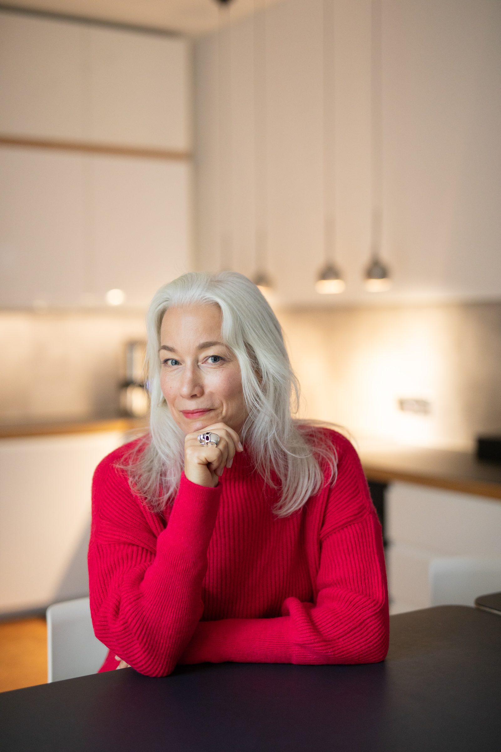 Nina Goldhammer in rotem Pullover sitzt an einem dunklen Tisch in einer hellen Küche und stützt das Kinn in die Hand