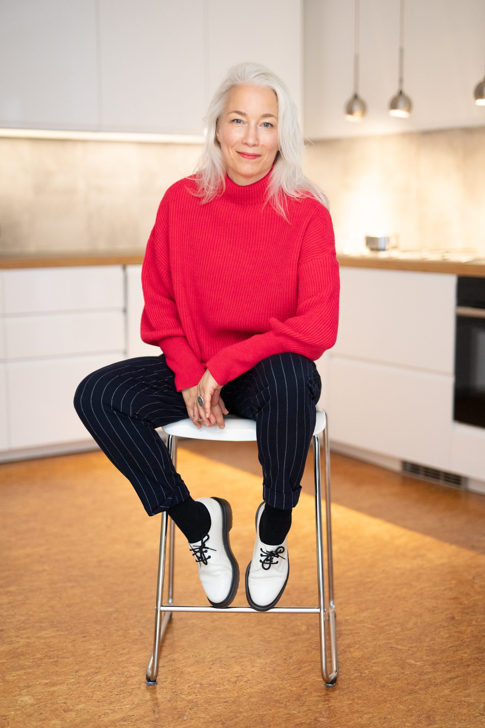 Nina Goldhammer in rotem Pullover, Nadelstreifenhose und weißen Schuhen sitzt auf einem Barhocker in der Küche