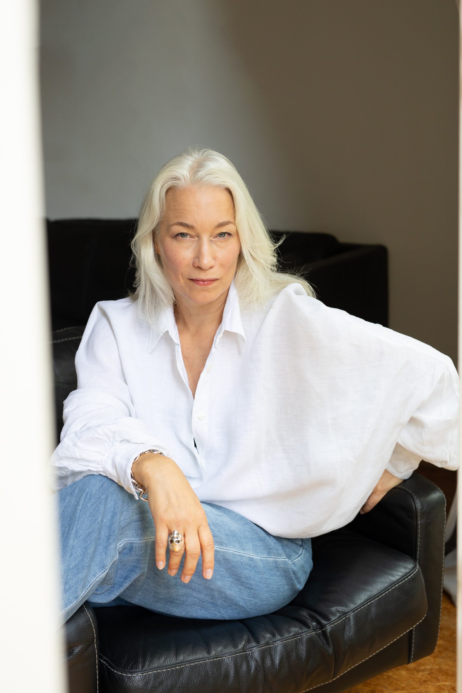Nina Goldhammer in weißer Bluse und Jeans sitzt in einem schwarzen Ledersessel, durch eine offene Tür fotografiert
