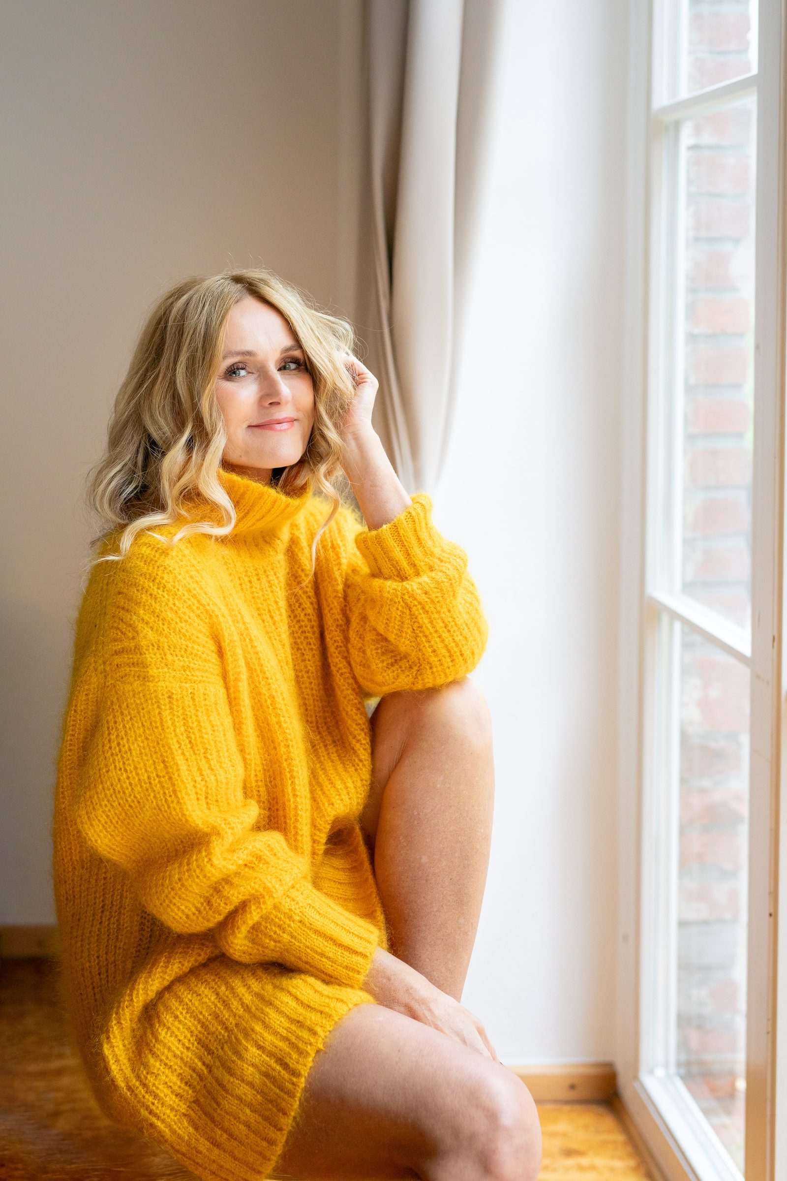 Nowshine in gelbem Strickpullover sitzt am Fenster und lächelt in die Kamera