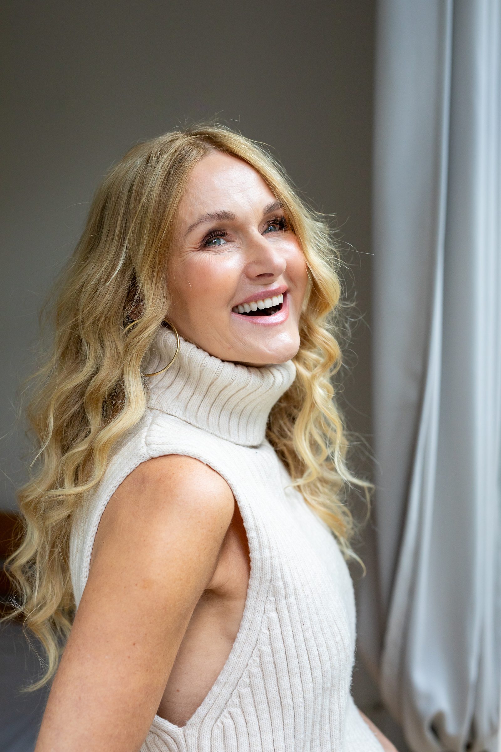 Nowshine im ärmellosen weißen Strickpullover lacht über die Schulter, daneben ein grauer Vorhang