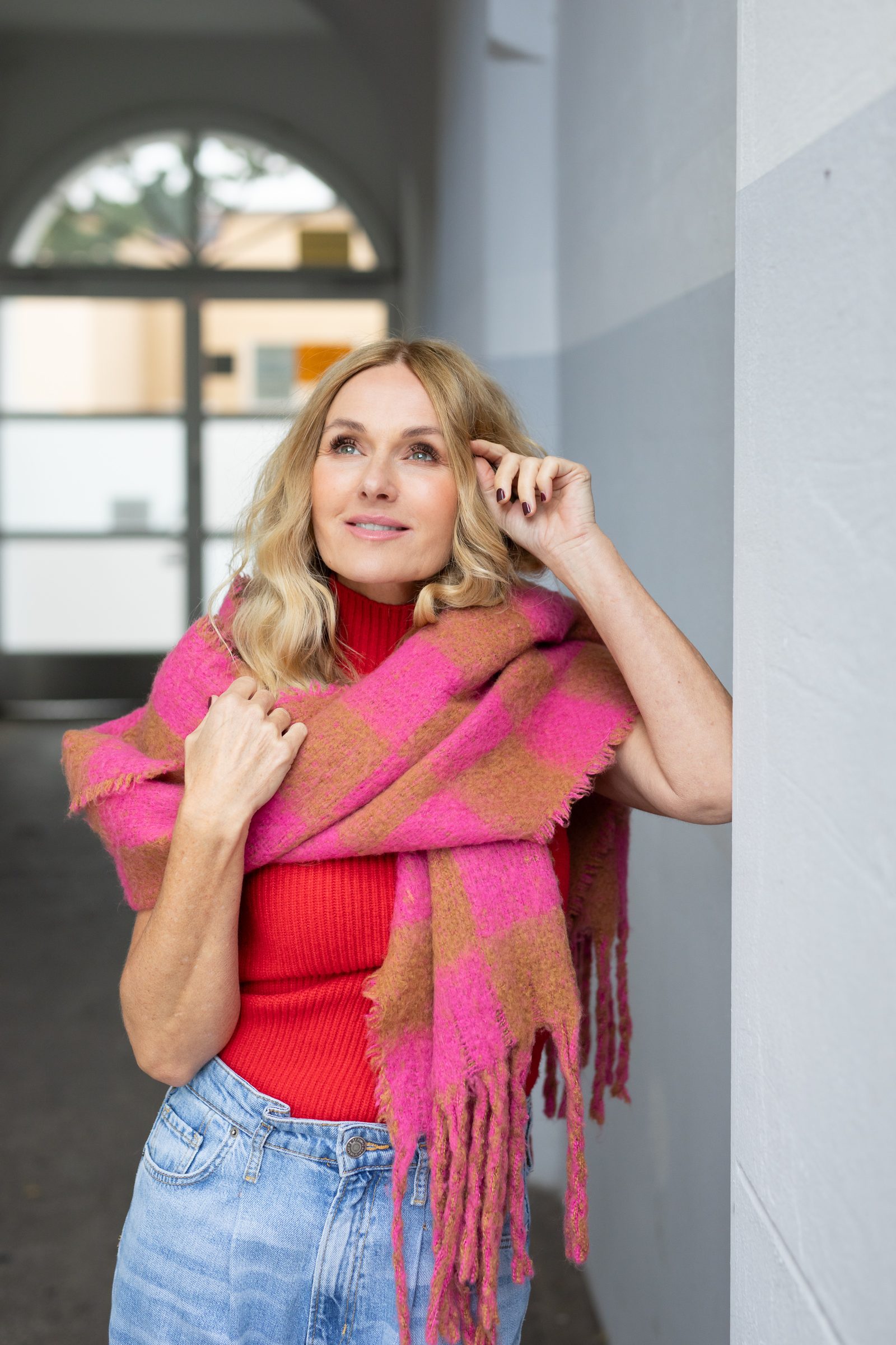 Nowshine mit pink und braun kariertem Schal lehnt an einer Wand im Flur und fasst sich ins Haar