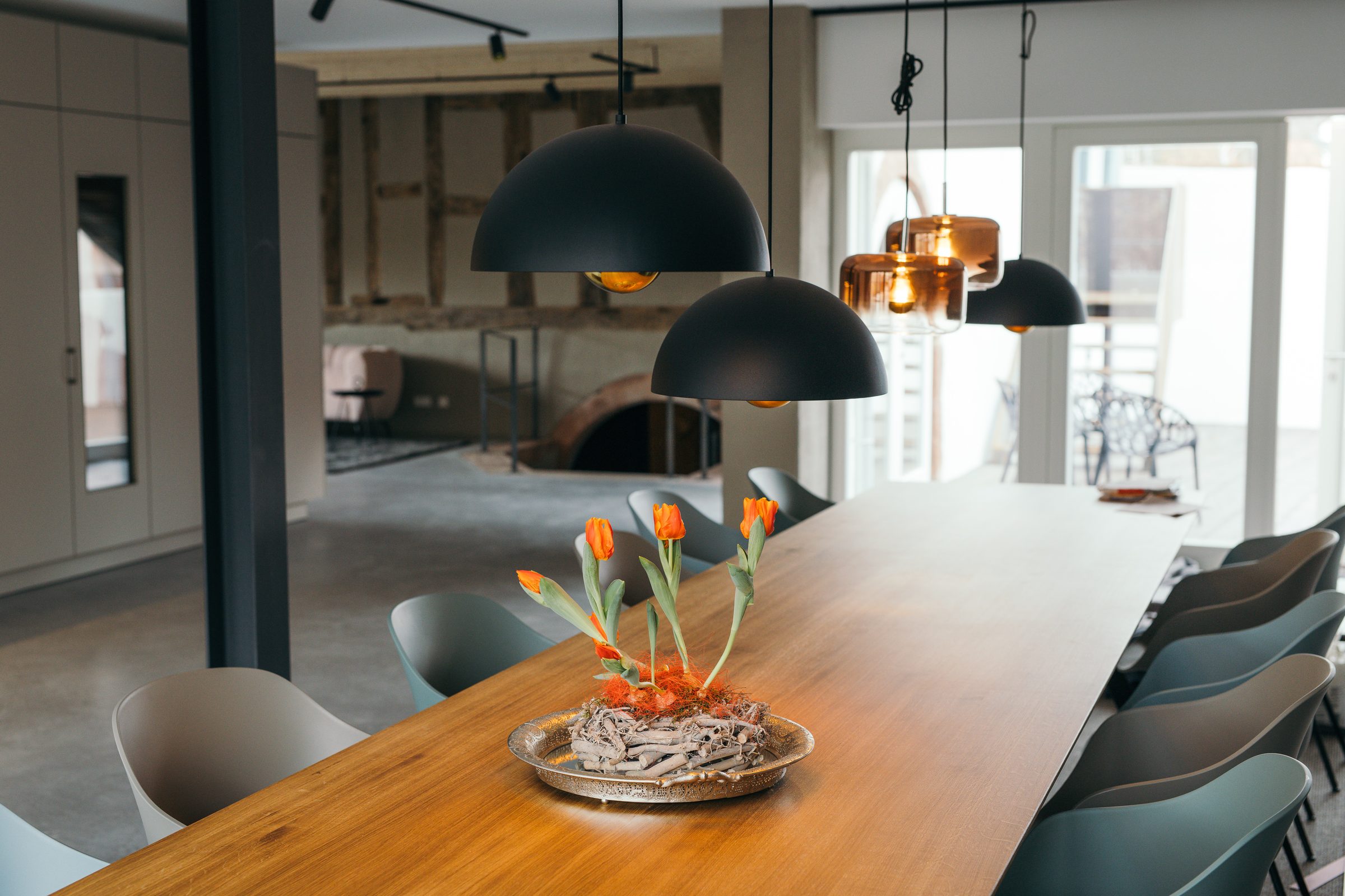 Lange Holztafel mit grauen Stühlen und Tulpengesteck unter schwarzen und kupferfarbenen Pendelleuchten im Ois is gut, dahinter altes Fachwerk