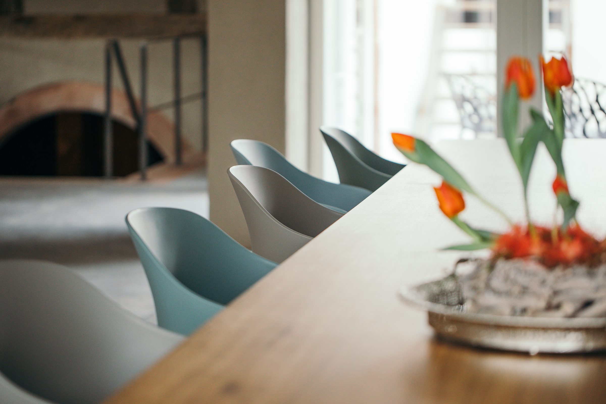 Ende der langen Holztafel mit grauen Stühlen und einem Gesteck aus orangefarbenen Tulpen, im Hintergrund ein alter Gewölbebogen