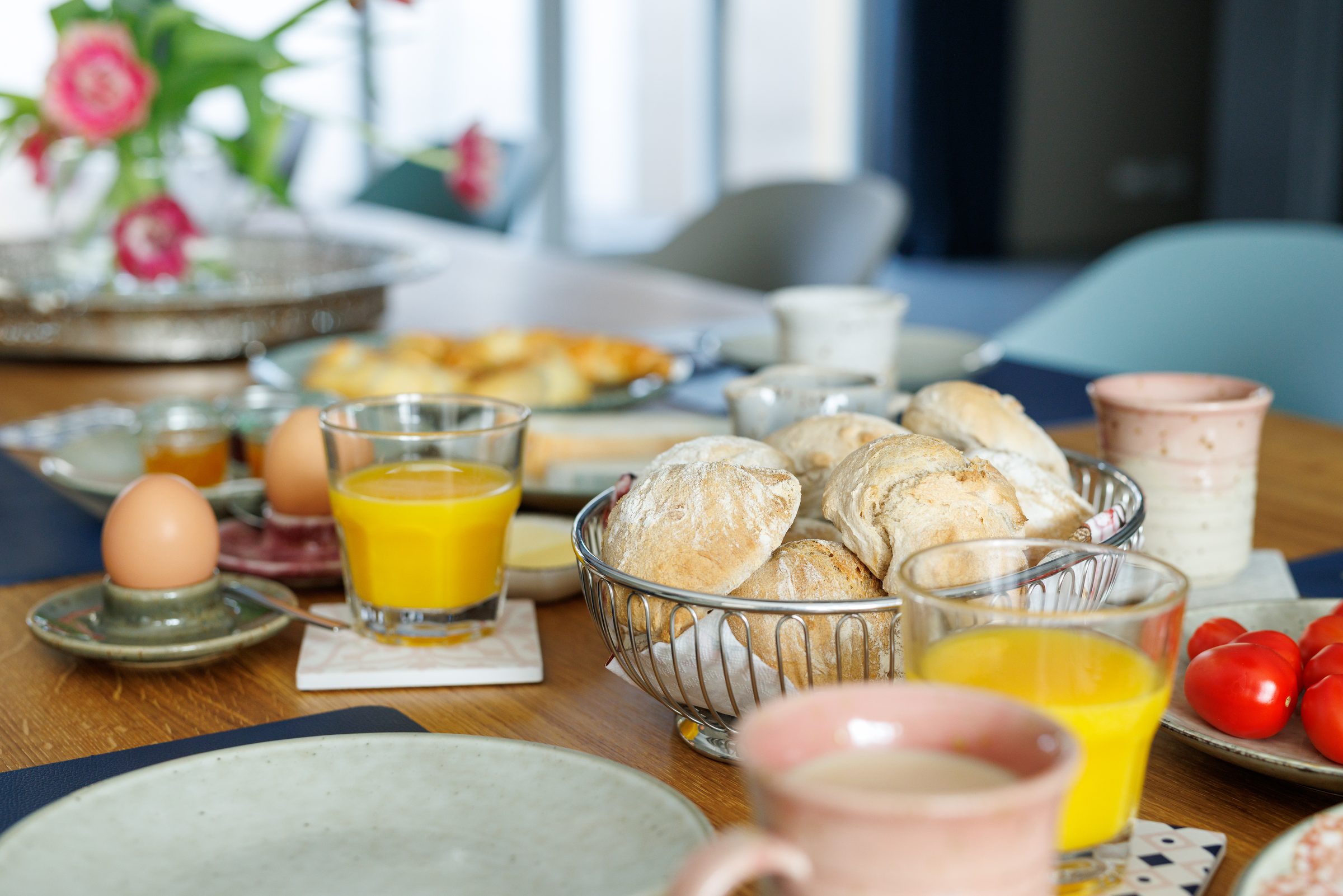 Frühstückstisch mit Brötchenkorb, Frühstückseiern, Orangensaft, Tomaten und Blumen