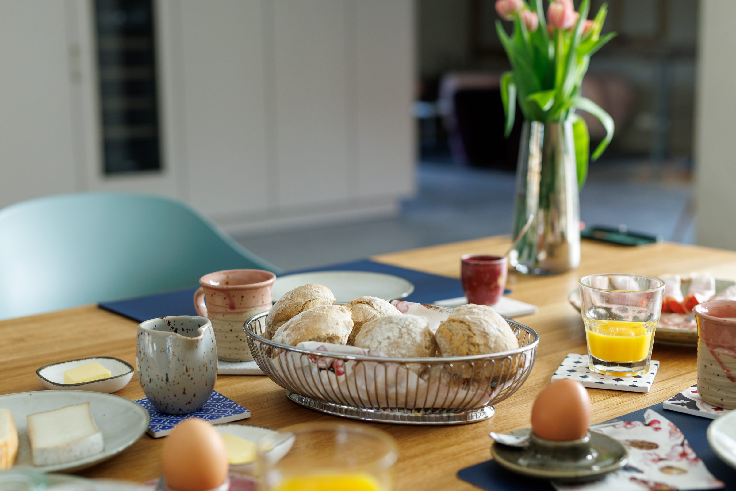 Frühstückstisch mit Brötchenkorb, Frühstücksei, Butter und Käse, dahinter Tulpen in einer Glasvase und graue Stühle