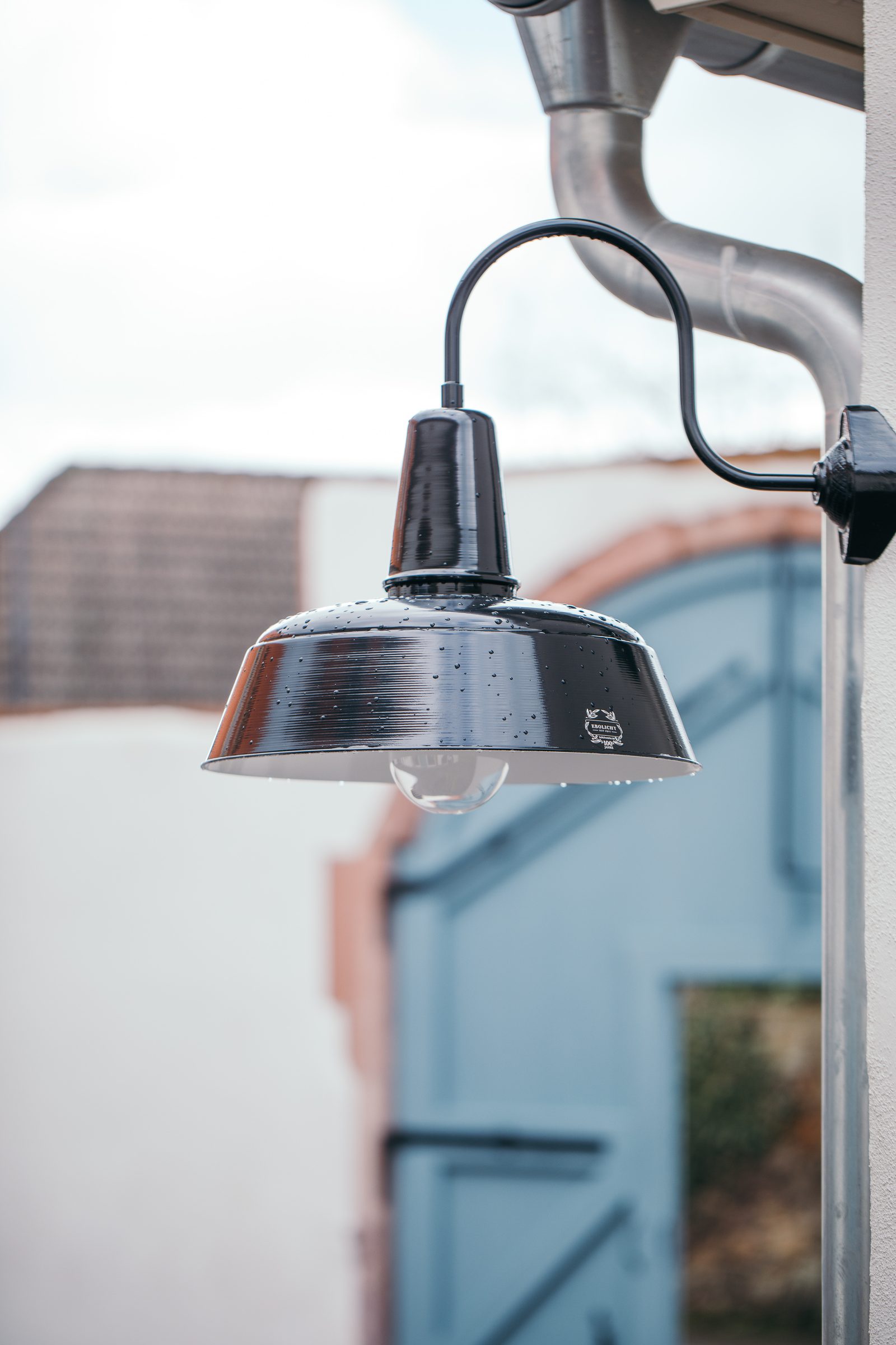 Schwarze Emaillelampe an einer Hauswand, dahinter unscharf ein hellblaues Tor im Sandsteinbogen