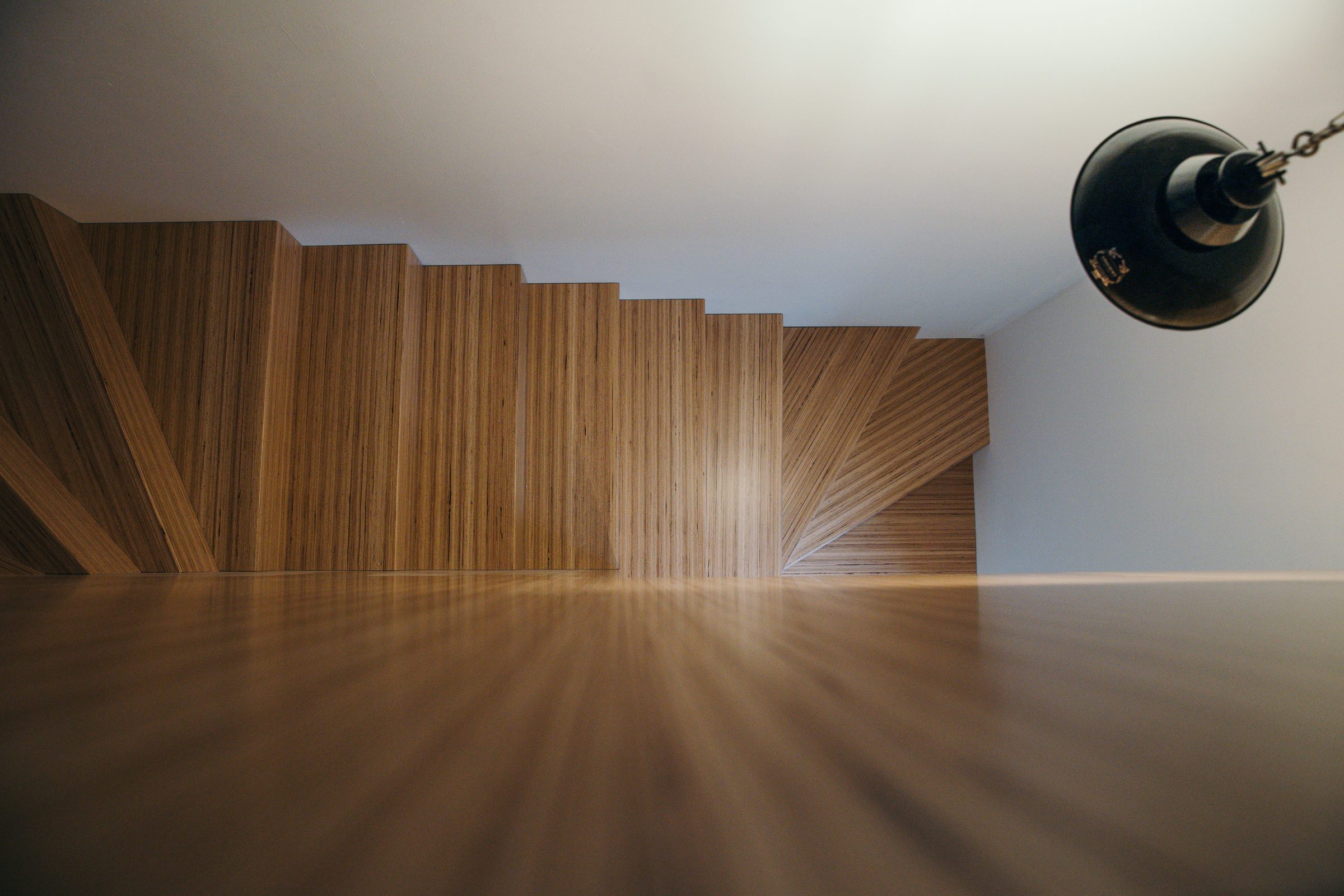 Treppe aus hellem Schichtholz von unten gesehen, vorne eine glänzende Holzfläche, rechts eine schwarze Hängelampe an einer Kette