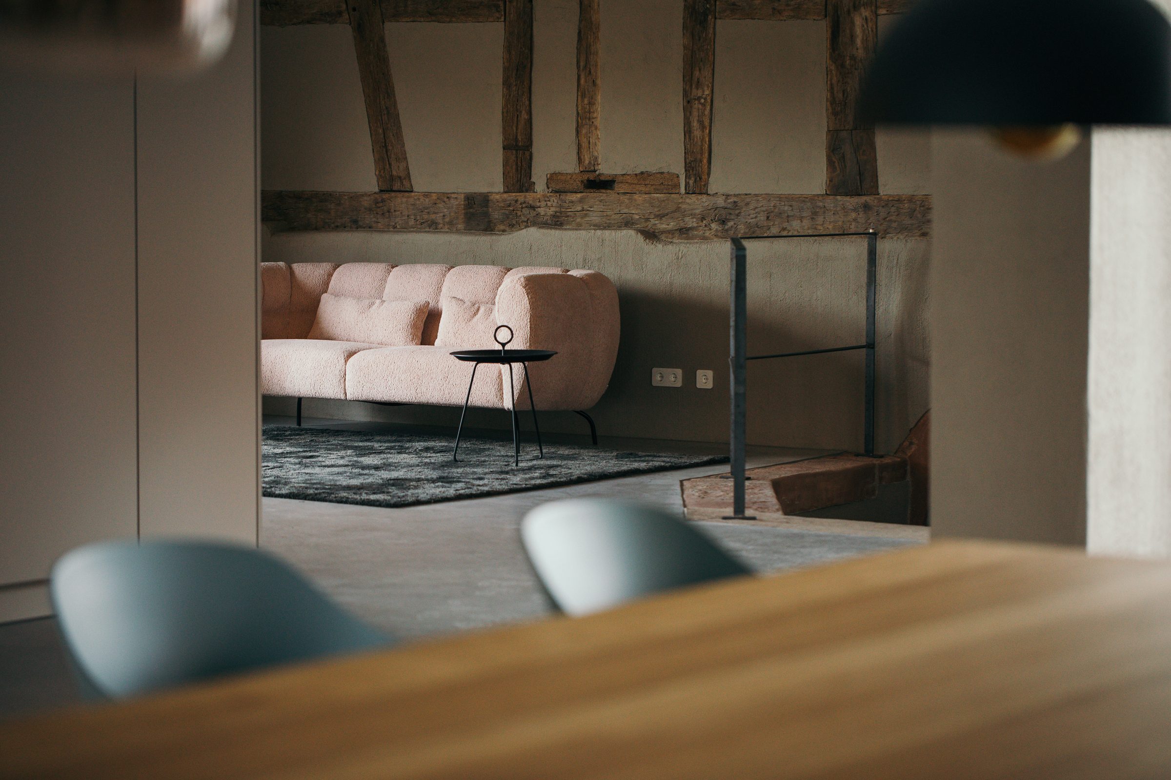 Rosa Sofa mit Beistelltisch hinter einer Fachwerkwand, im Vordergrund die Holztafel mit grauen Stühlen