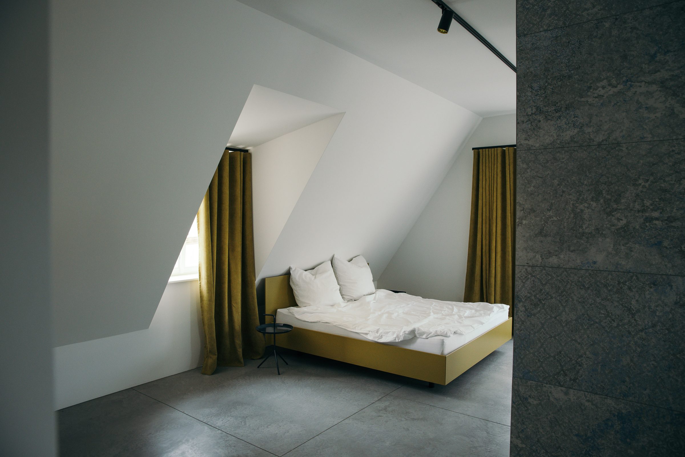 Doppelbett mit senfgelbem Rahmen unter einer Dachschräge im Ois is gut, an den Gauben goldgelbe Vorhänge, rechts eine dunkle Steinwand