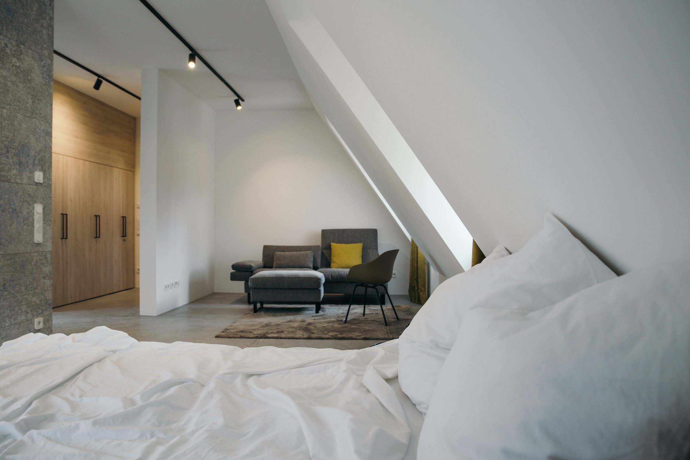 Blick vom Bett über den Betonboden zu einer Sitzecke mit grauem Sofa und gelbem Kissen unter der Dachschräge